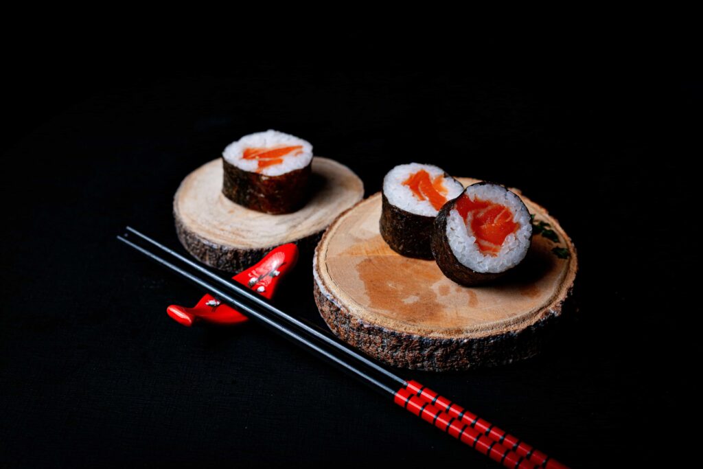 An aesthetically photographed sushi on a wooden platter accompanied by black chopsticks with a black background.