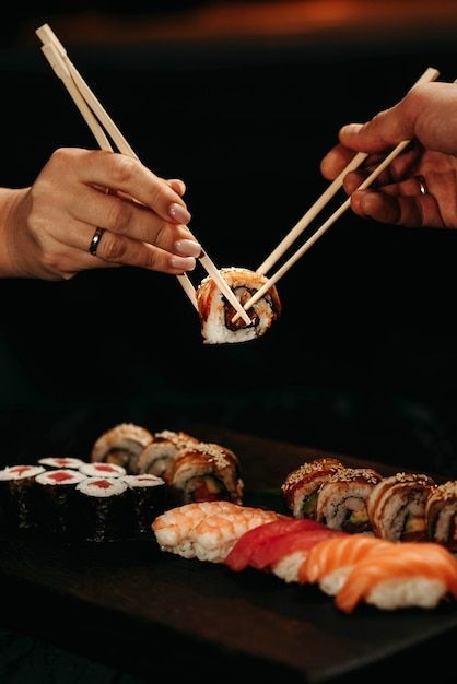 Two people picking up sushi rolls with chopsticks from a wooden platter of assorted sushi.