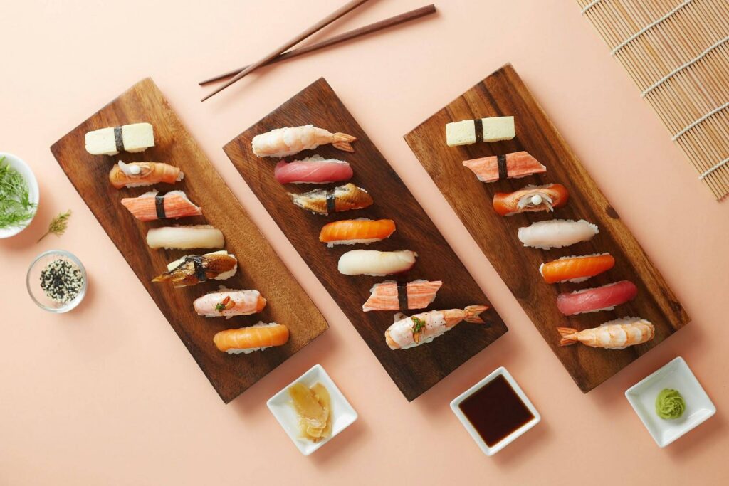 Overhead shot of three dark wooden serving boards displaying assorted sushi and nigiri, including ebi (shrimp), maguro (tuna), tamagoyaki (egg), and crab stick, set with ginger, soy sauce, and wasabi.