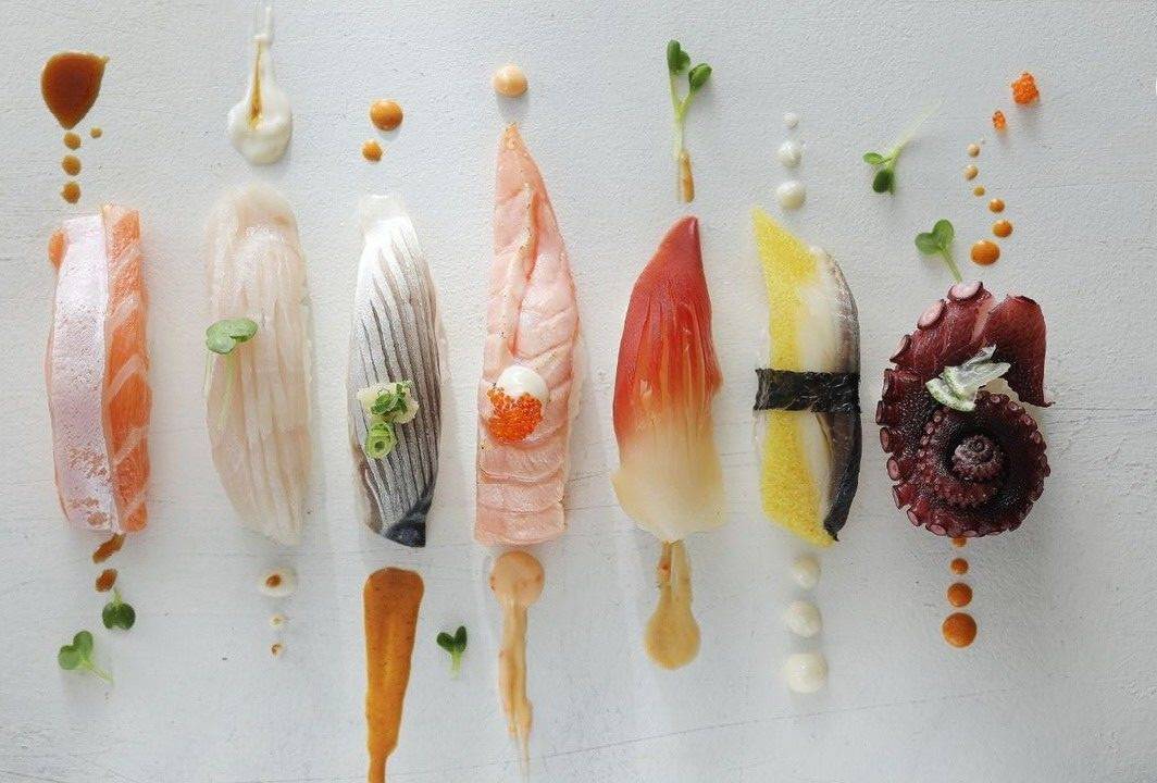 Artistic presentation of diverse nigiri and sashimi cuts, including salmon, snapper, mackerel, clam, and octopus, each expertly dressed with sauces and microgreens on a white background.