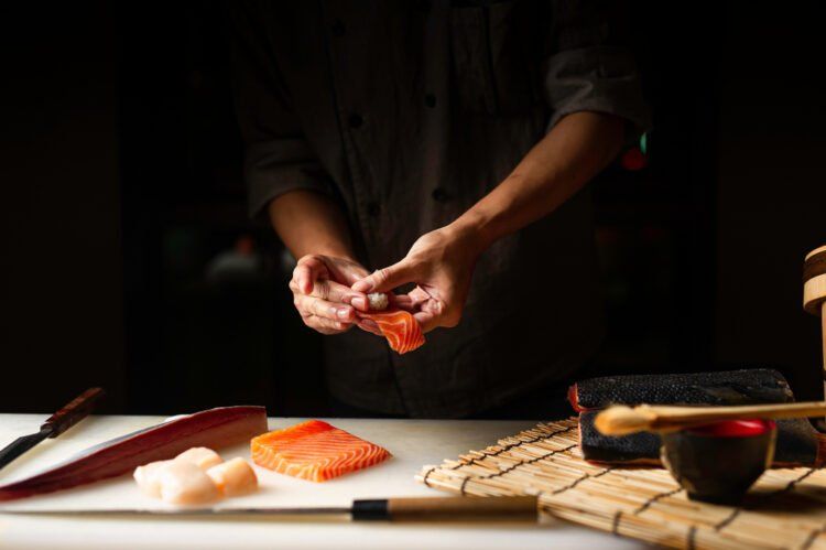 Close-up of a sushi chef's hands meticulously forming a piece of nigiri with fresh salmon and seafood on a white cutting board, emphasizing the artisanal process.