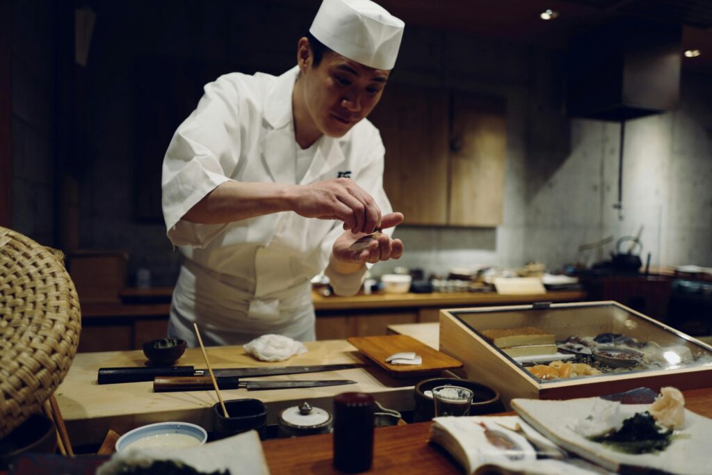 In a dimly lit, traditional setting, a focused sushi chef wearing a white uniform and hat carefully shapes a piece of sushi with his hands. The wooden counter in front of him displays culinary tools such as knives and a box of fresh ingredients, emphasizing the craftsmanship involved in the preparation.