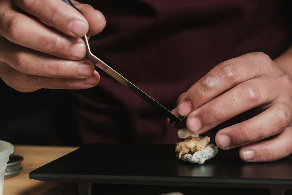 This extreme close-up highlights the meticulous precision of a chef using metal tweezers to place a delicate garnish, likely caviar, onto a small, intricate appetizer. The focus remains tightly on the hands and the culinary creation resting on a matte black plate, emphasizing the artistry and attention to detail involved in gourmet food plating.