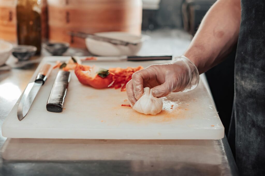 This close-up captures a chef's gloved hand holding a bulb of fresh garlic on a white cutting board, surrounded by slices of red bell pepper. A sharp knife lies ready nearby, emphasizing the active and methodical process of ingredient preparation in a kitchen setting.