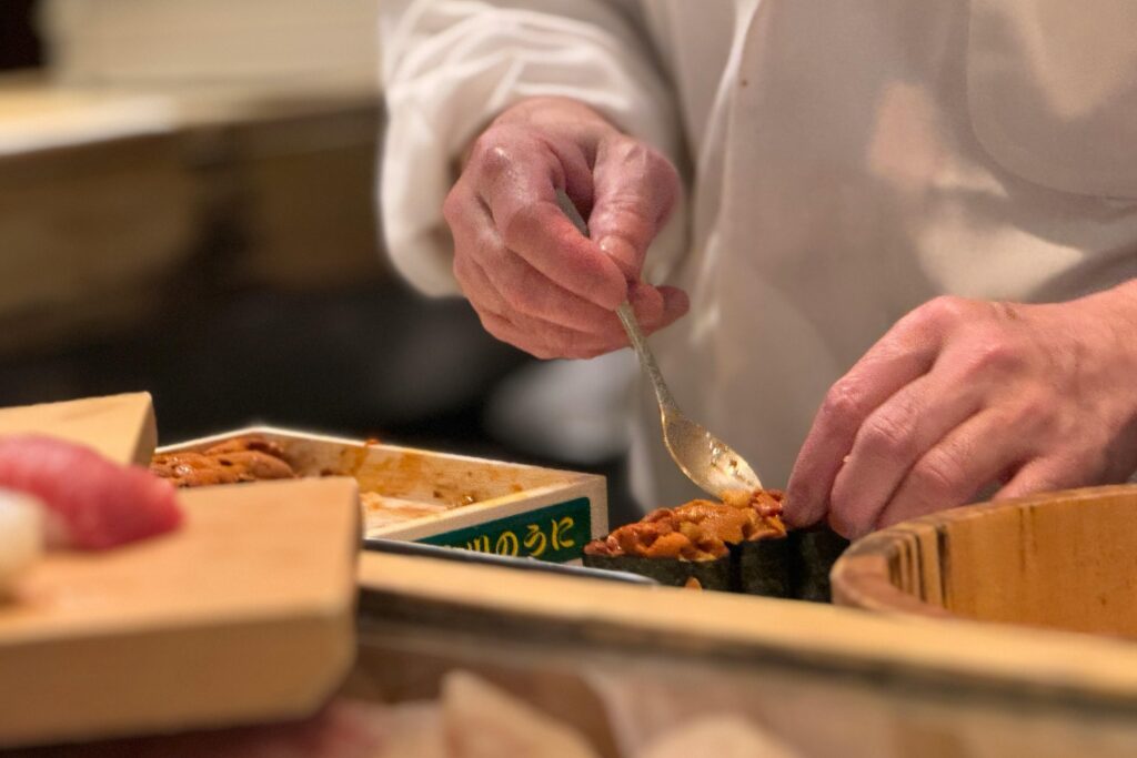 This close-up captures the delicate hands of a sushi chef using a small spoon to carefully top a piece of gunkan-maki with fresh, vibrant orange sea urchin. Surrounding the preparation area are wooden trays of ingredients and other sushi pieces, emphasizing the freshness and artisanal quality of the meal.