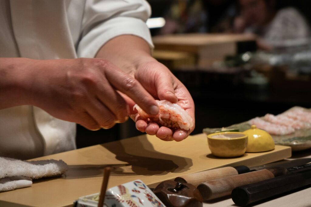 This close-up photograph captures the skilled hands of a sushi chef delicately shaping a piece of nigiri topped with raw shrimp. In the foreground, traditional wooden-handled knives and small ceramic dishes sit on the counter, while the blurred background suggests a busy dining environment.