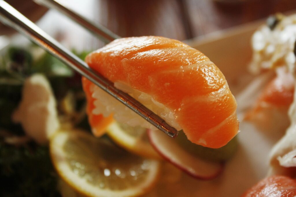 A pair of metal chopsticks holds a piece of salmon nigiri, showcasing the glossy, vibrant orange fish draped over a small mound of white rice. The background is softly blurred, revealing lemon slices and greens on a plate that suggest a fresh and appetizing dining setting.