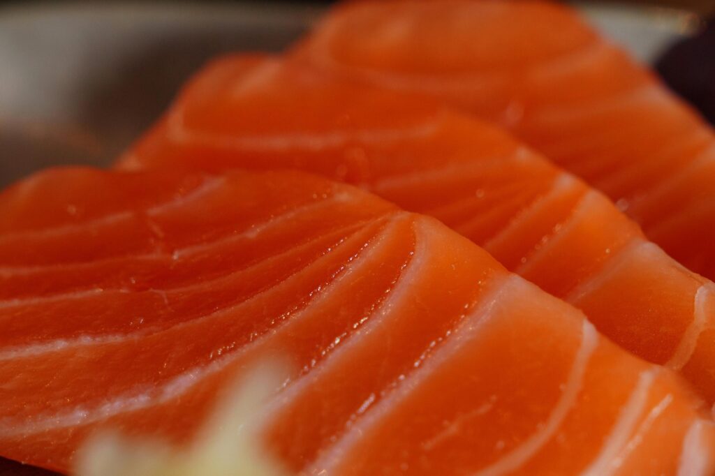 This close-up image captures the rich texture of fresh salmon slices, featuring vibrant orange flesh striated with distinct, curved white lines of fat. The overlapping arrangement and glossy sheen emphasize the succulent and high-quality nature of the sashimi.