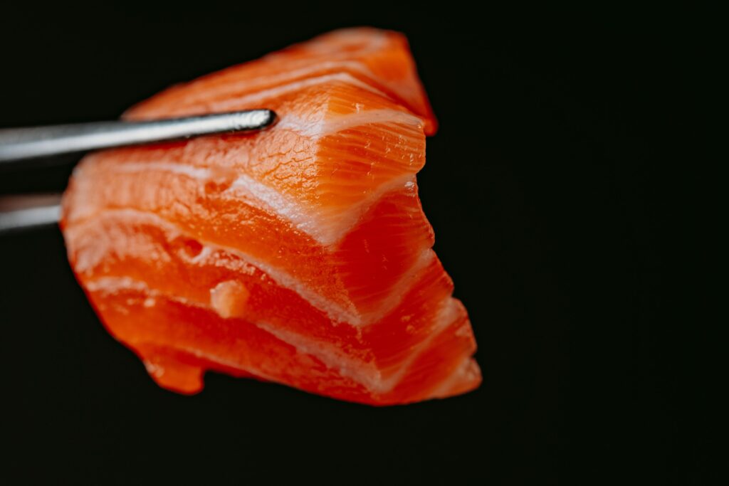 Against a solid black background, silver tweezers hold a thick, vibrant slice of salmon, creating a striking contrast. The close-up view emphasizes the fish's glossy surface and the prominent white lines of fat running through the rich orange flesh.