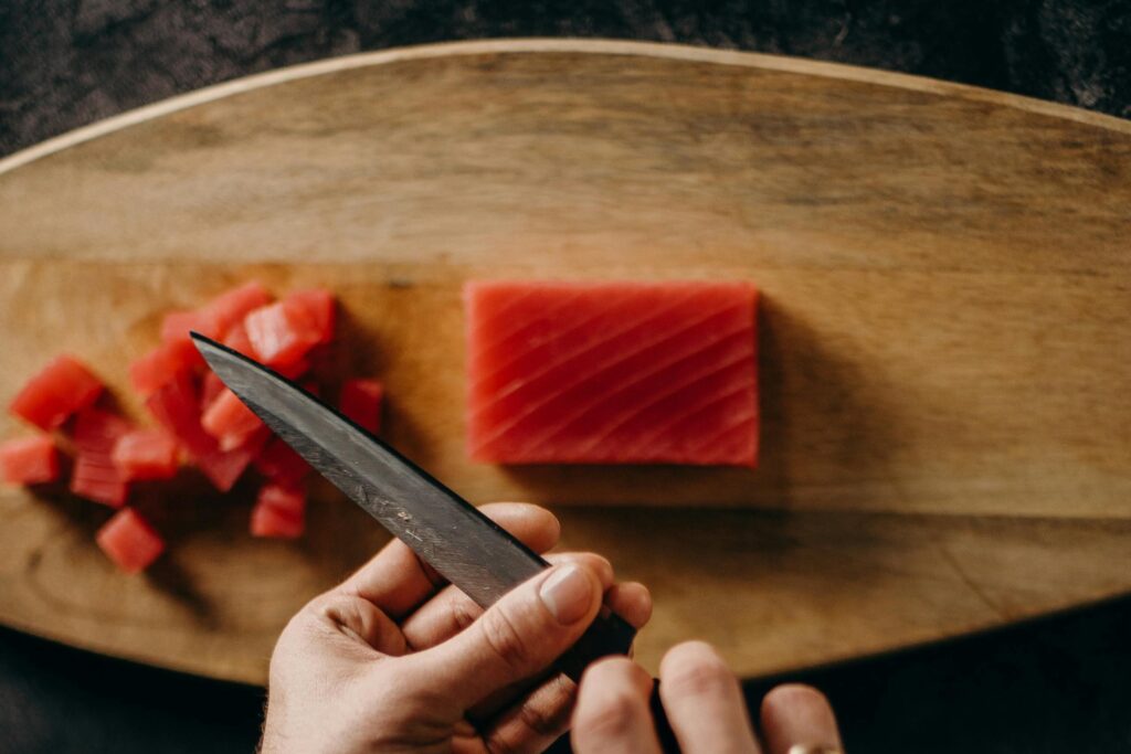 From a top-down perspective, a pair of hands holds a knife over a wooden cutting board containing a mound of diced red fish and a rectangular block of the same fillet. The scene captures a moment in culinary preparation, contrasting the finished cubed portions with the remaining uncut meat.