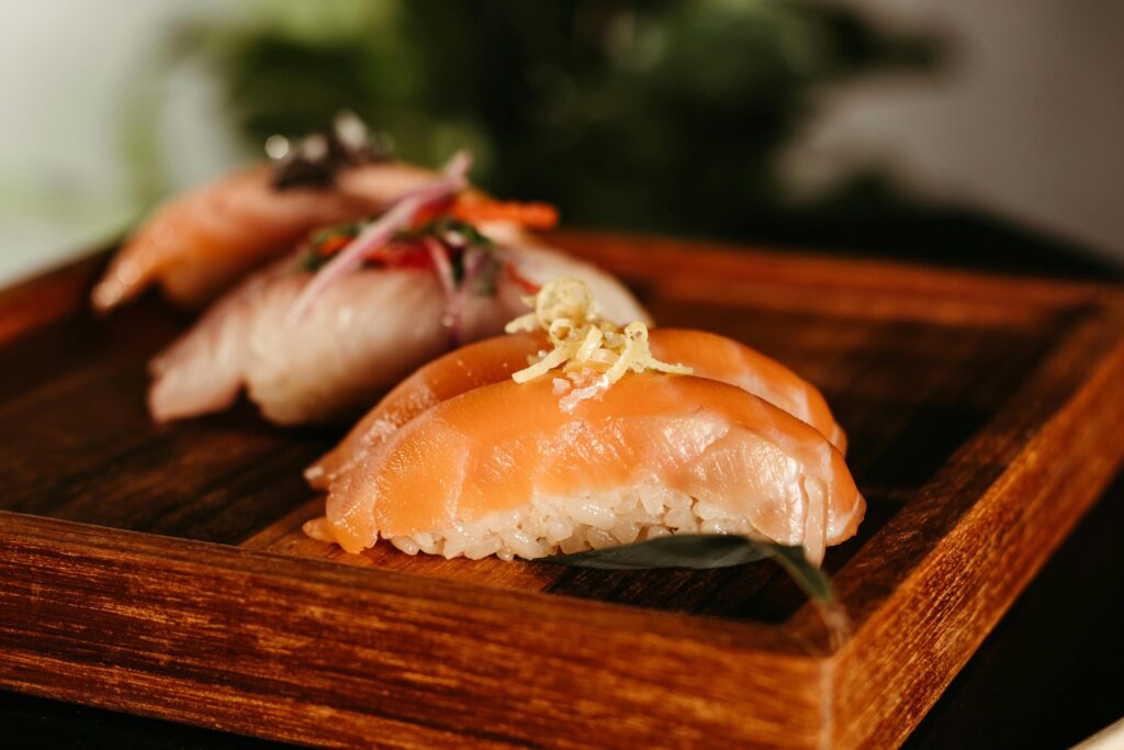 This close-up photograph highlights two pieces of fresh salmon nigiri topped with delicate zest, sitting prominently on a rustic wooden tray. The shallow depth of field softens the appearance of the third piece of sushi in the background.