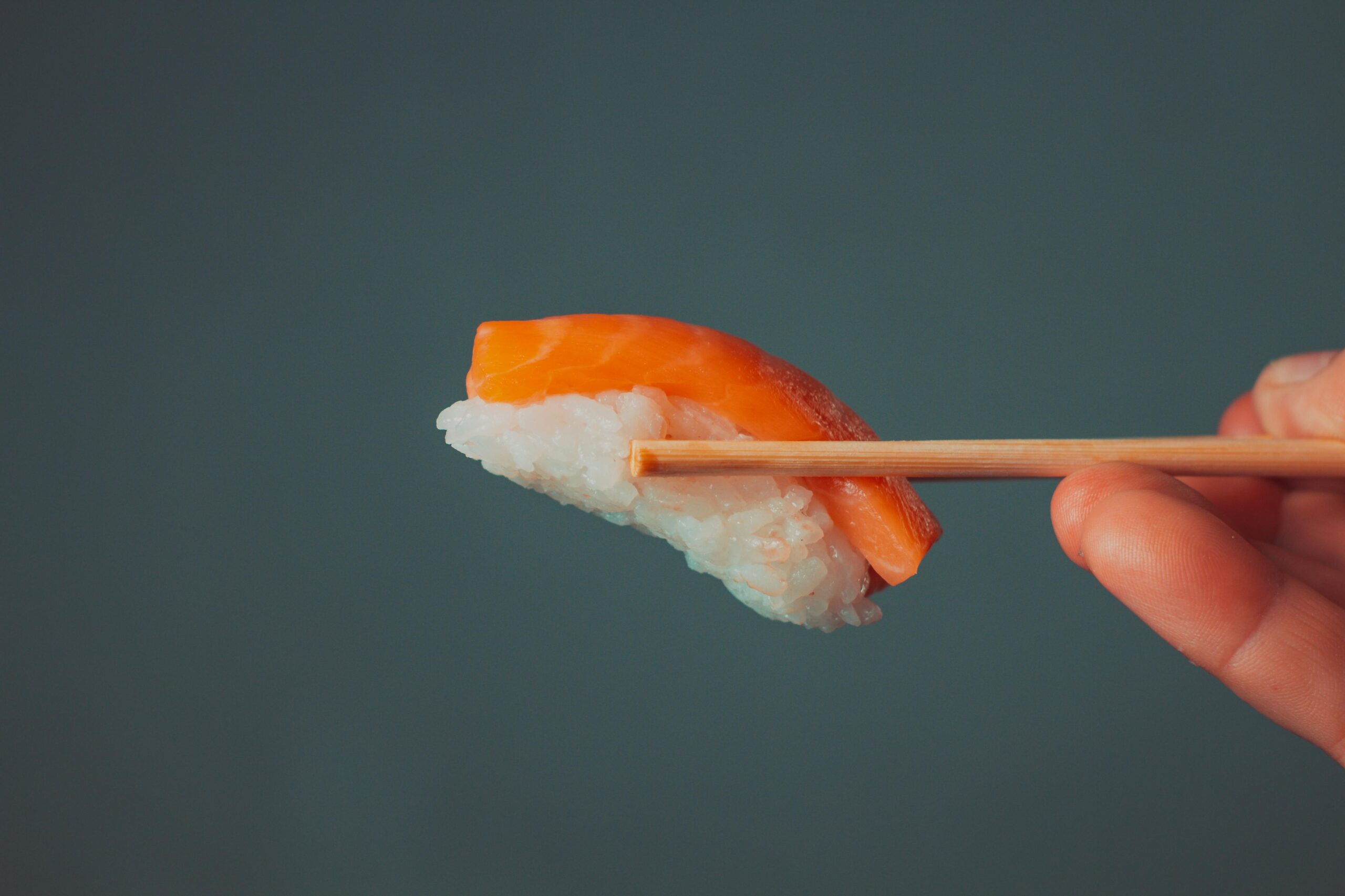 This minimalist photograph captures a single piece of salmon nigiri held by wooden chopsticks against a plain, dark grey background. The side-view composition draws attention to the vibrant orange salmon and the texture of the white rice, emphasizing the simplicity of the sushi.