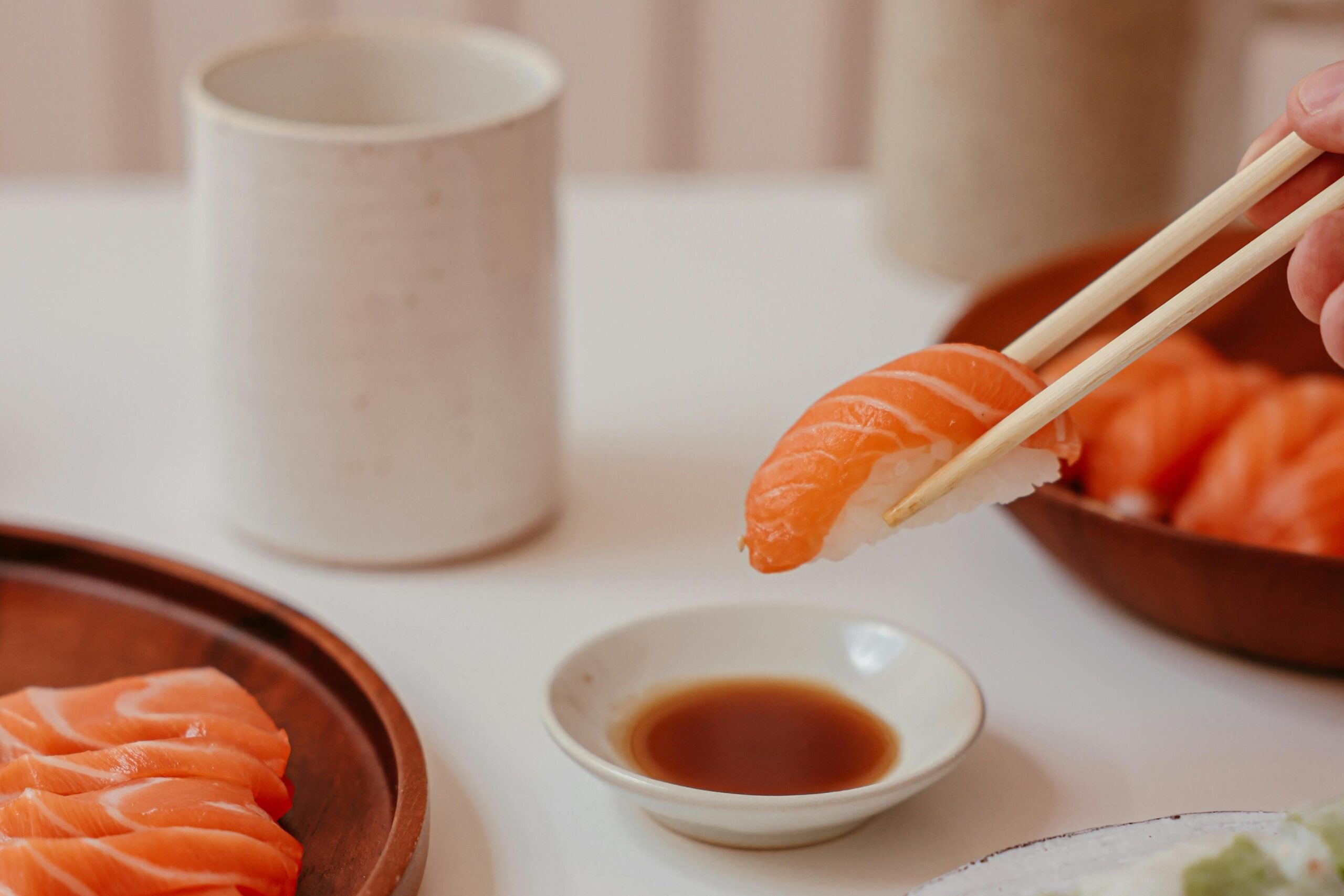 This close-up shot depicts a piece of salmon nigiri held by wooden chopsticks, hovering just above a small dish of soy sauce. In the soft-focus background, wooden plates filled with fresh salmon sashimi and a ceramic cup rest on a white table.