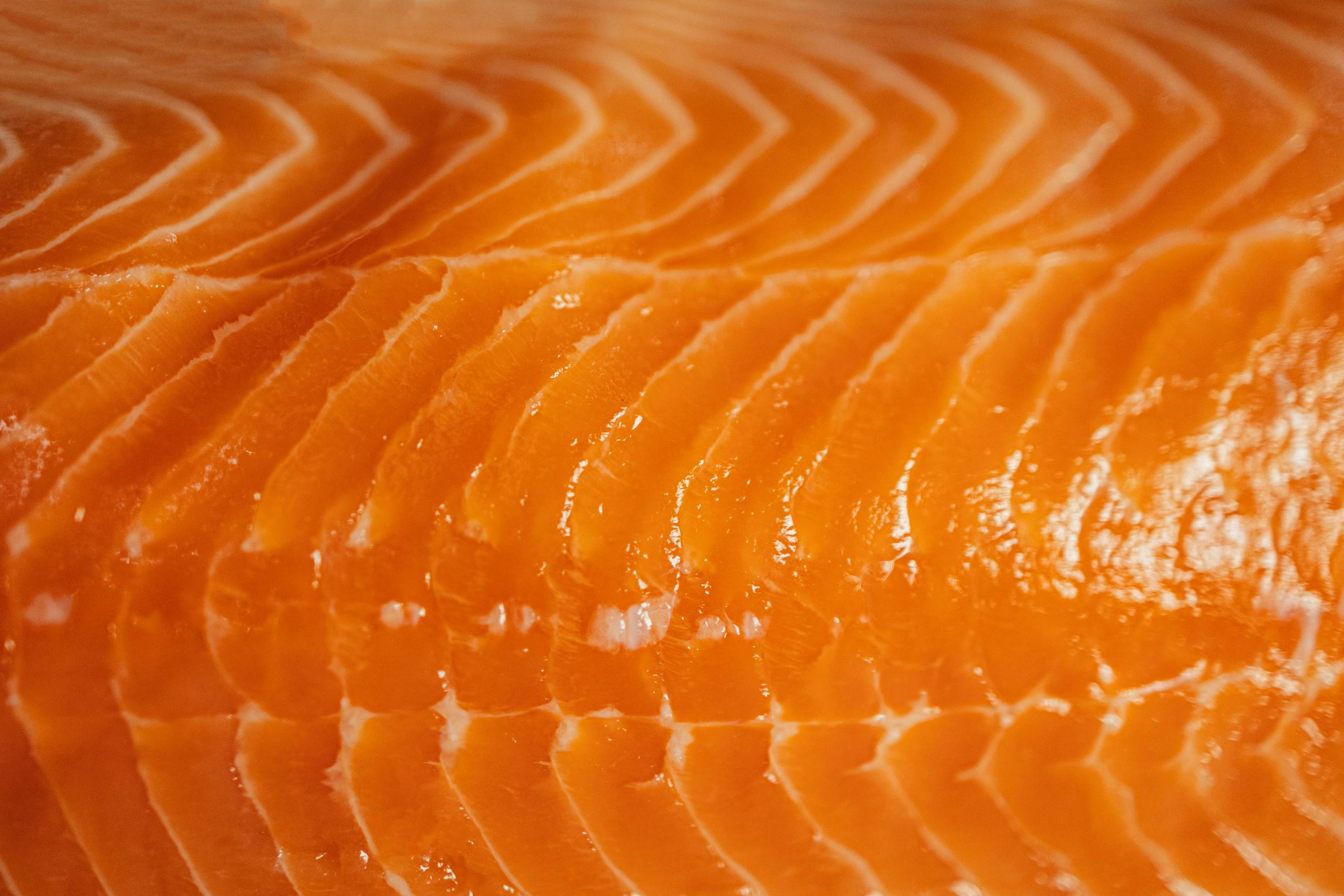 This extreme close-up captures the rich, vibrant orange hues of a raw salmon fillet, emphasizing the glossy and moist texture of the meat. Distinct white striations of fat ripple across the surface in a repetitive wave pattern, highlighting the detailed marbling of the fresh fish.