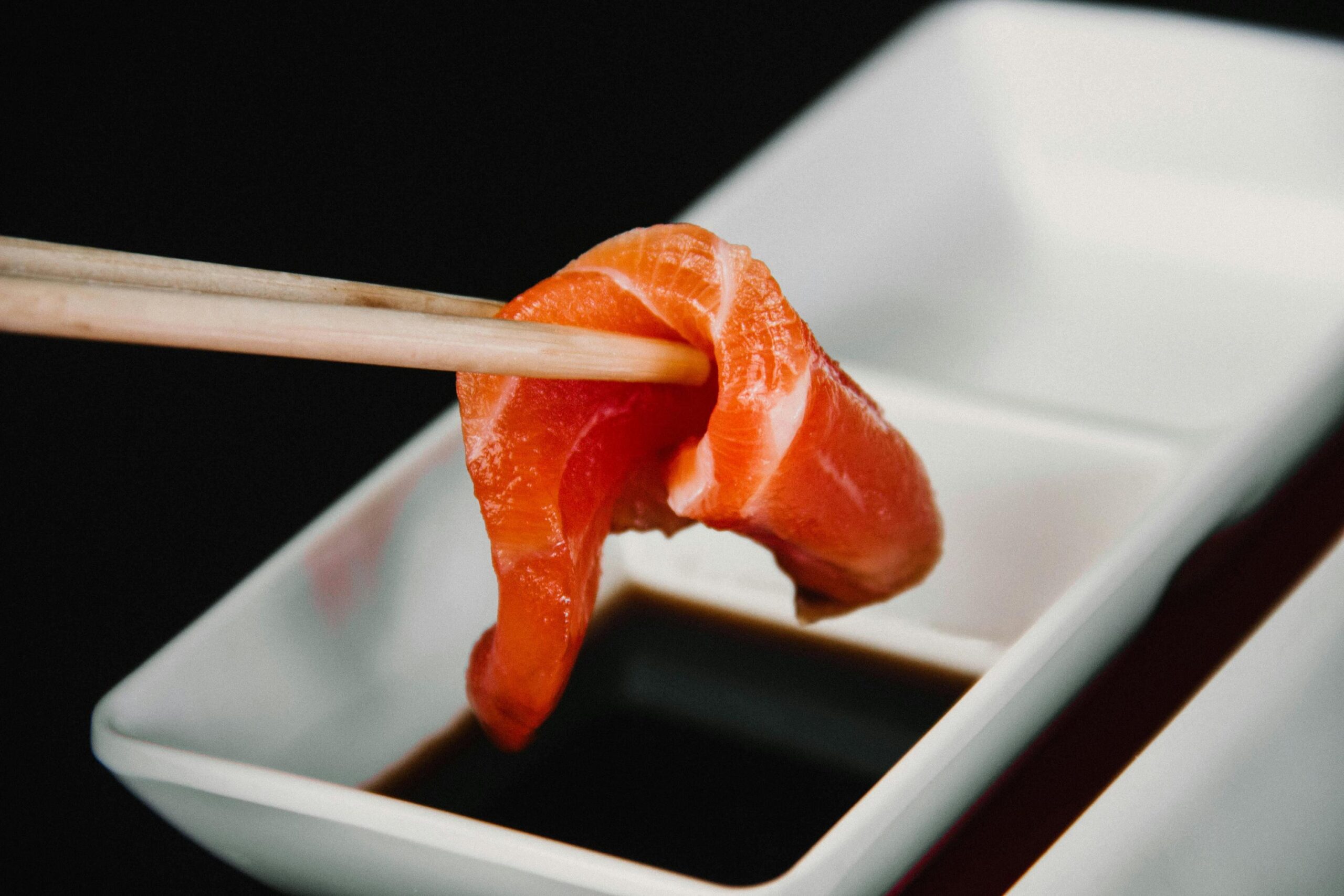 This close-up image features a slice of fresh salmon sashimi draped over wooden chopsticks, hovering just above a small white rectangular dish filled with dark soy sauce. The vibrant orange flesh of the fish contrasts starkly against the dark background.