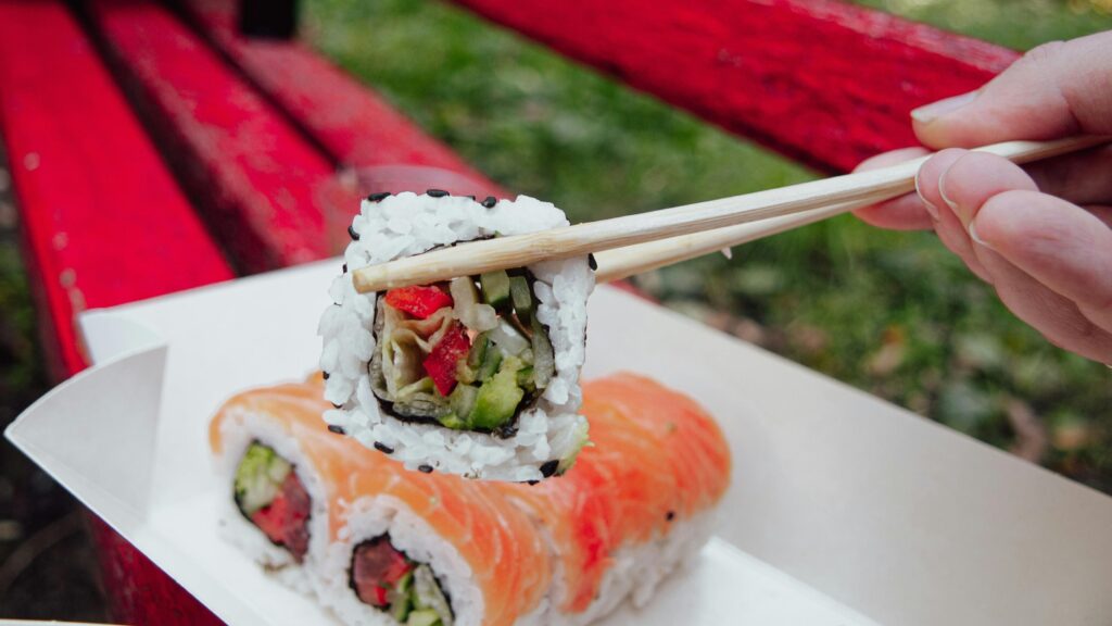 This image features a hand holding a piece of sushi with chopsticks over a paper take-out tray containing two other salmon-wrapped rolls.