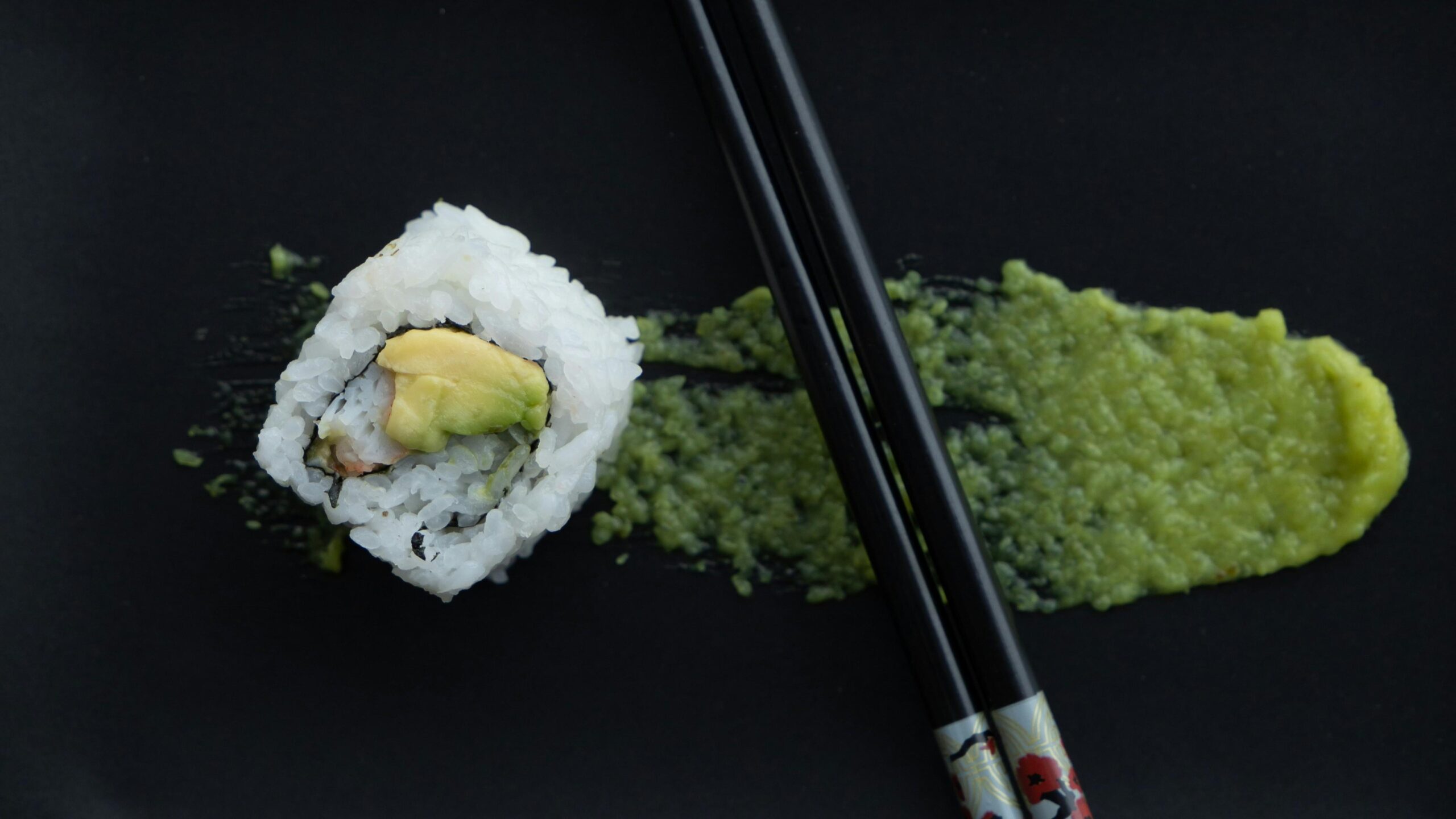 This overhead shot features a single piece of sushi roll topped with avocado, resting on a matte black plate. A dark pair of chopsticks lies diagonally across a smear of bright green wasabi.