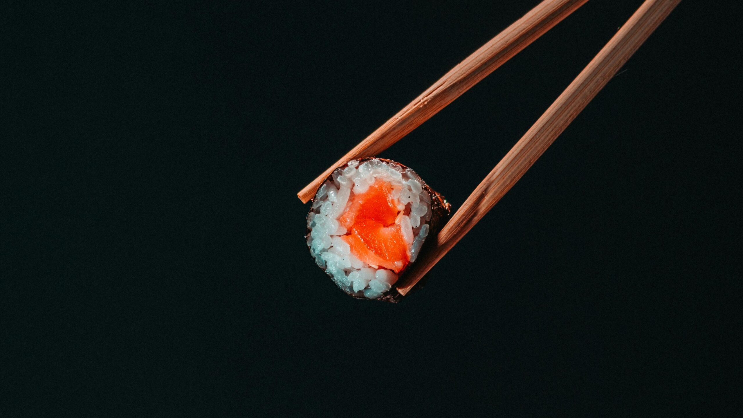 This is a minimalist, close-up shot of a single piece of salmon sushi being held by wooden chopsticks against a pure black background.