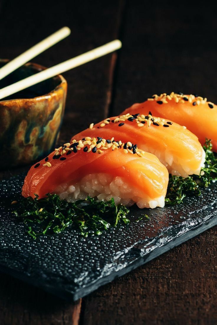 Close-up of a premium salmon and avocado sushi roll being held by wooden chopsticks and dipped into a small, elegant bowl of soy sauce.