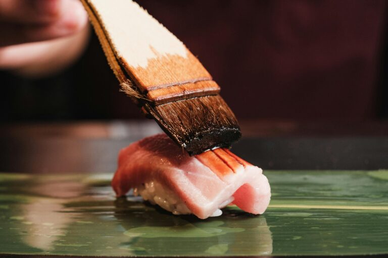 This extreme close-up captures a chef delicately brushing a dark glaze onto a piece of premium fatty tuna sushi resting on a green leaf. The glistening sauce highlights the rich marbling of the pink fish, emphasizing the precision and artistry involved in high-end culinary preparation.