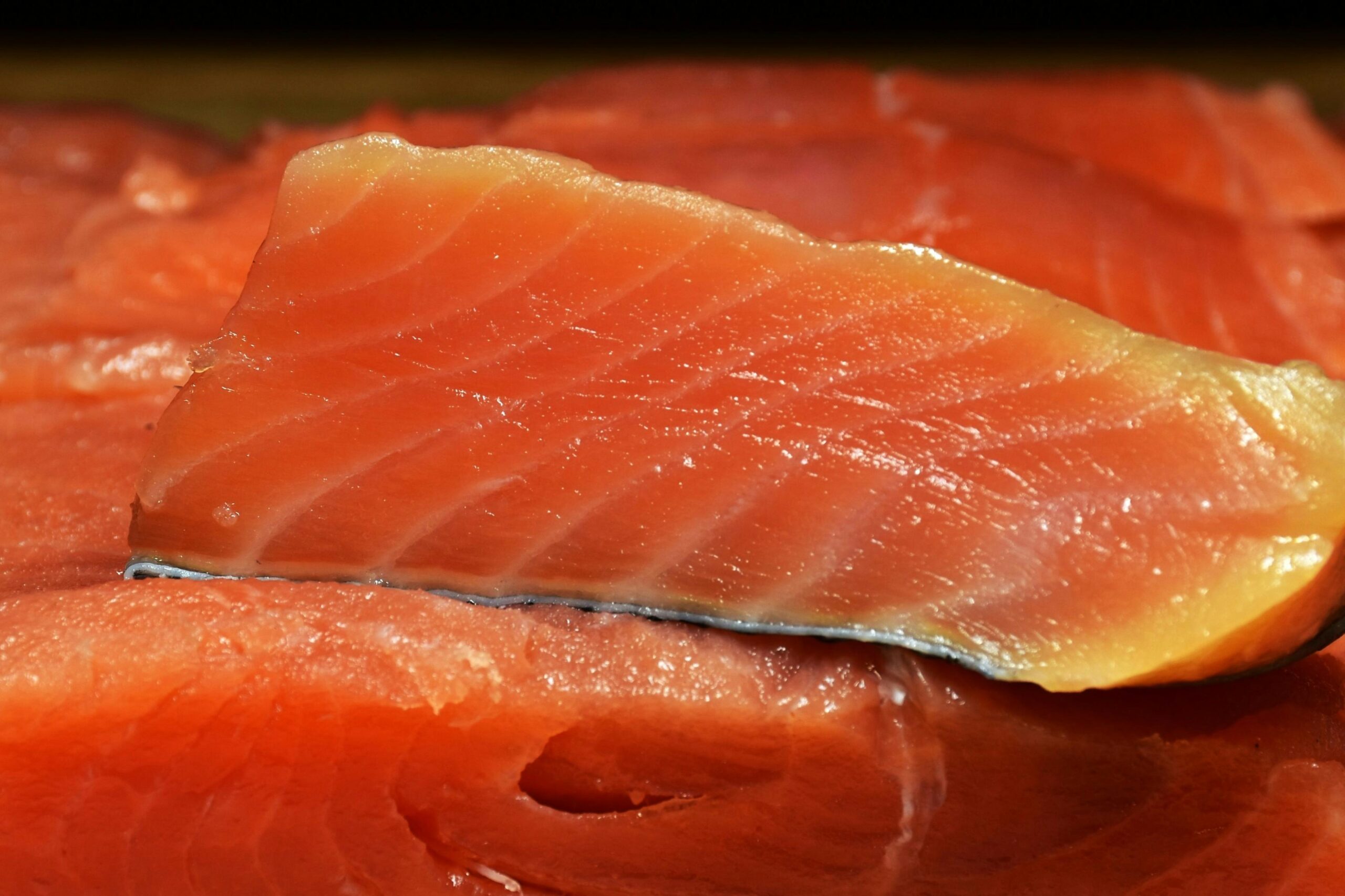 This close-up image features a slice of vibrant orange salmon, highlighting its glistening texture and distinct white lines of fat known as myomeres. The fillet rests against a background of similar fish, showcasing the moist quality of the seafood and the sliver of silver skin along the bottom edge.