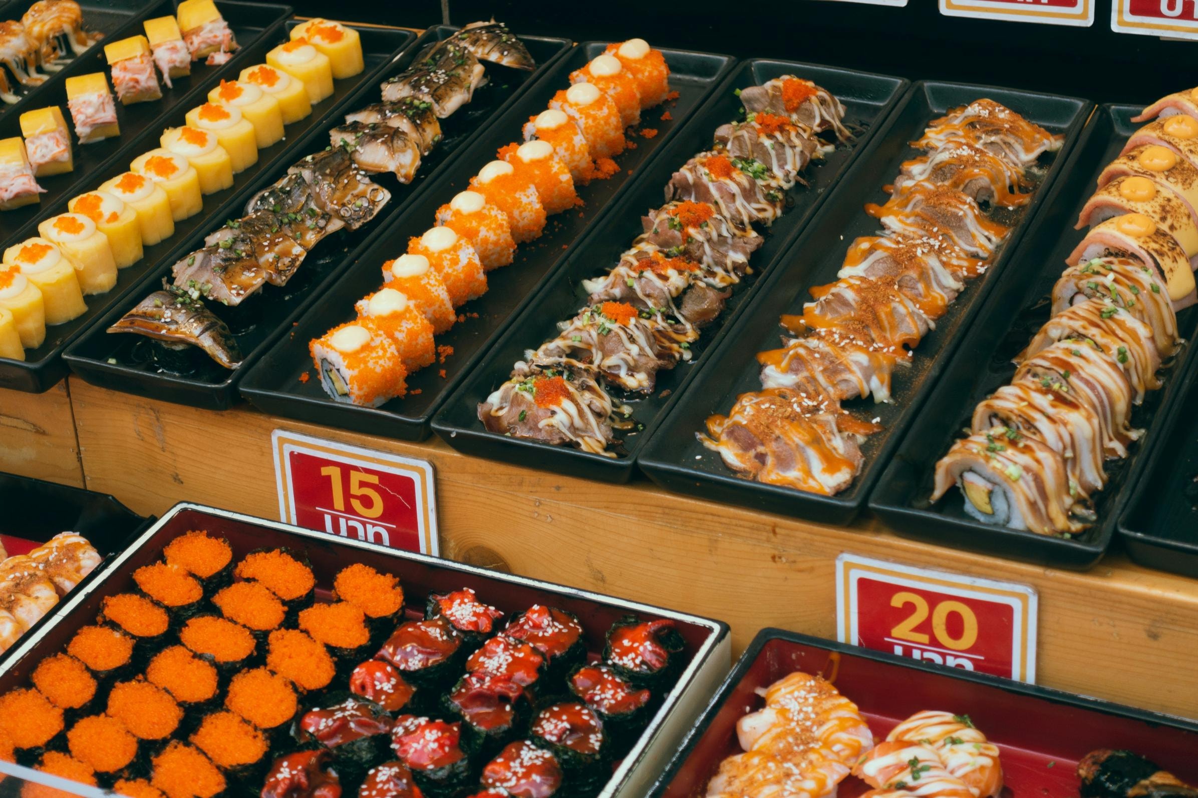 This photo showcases a vibrant display of various sushi types arranged in neat rows on black trays, including roe-covered rolls, seared nigiri with drizzled sauces, and gunkan maki. The organized presentation on wooden shelves, accompanied by visible price tags, suggests a bustling market or food stall offering a wide selection of Japanese delicacies.