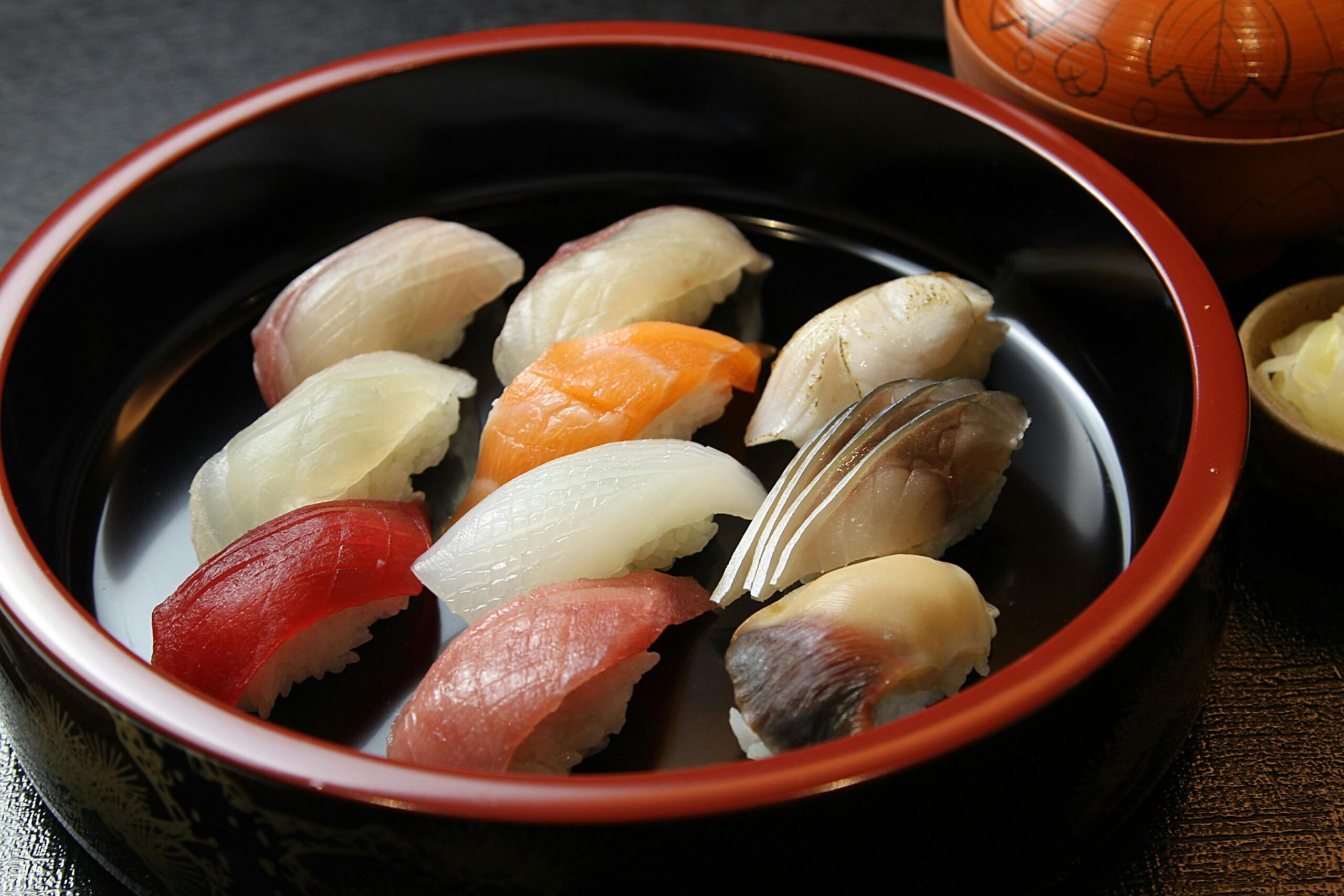 This image features a traditional round lacquerware bowl elegantly arranged with an assortment of fresh nigiri sushi, showcasing varieties like tuna, salmon, squid, and mackerel. To the side, a small dish of pickled ginger and a covered wooden bowl complement the main course, suggesting a complete and authentic Japanese dining experience.