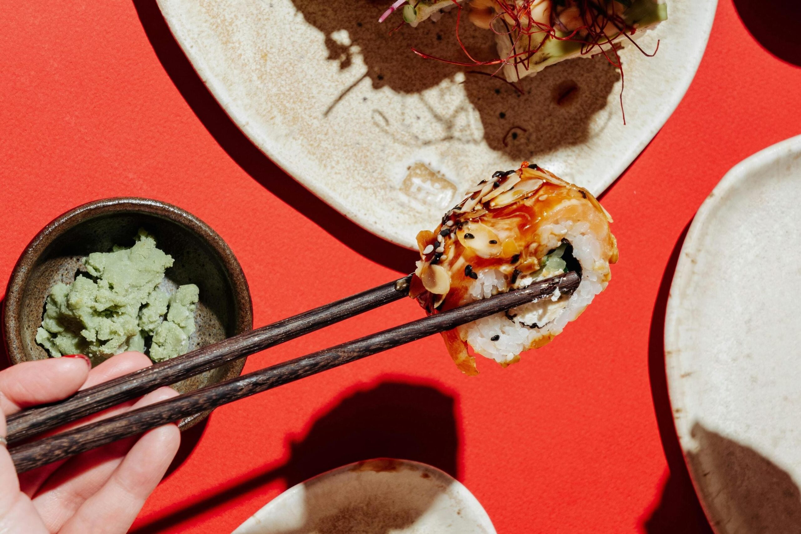 A hand holds dark wooden chopsticks gripping a sushi roll topped with almond flakes and savory glaze, set against a vivid red background. To the side, a small bowl of green wasabi paste accompanies the dish, while the overhead lighting casts sharp, dramatic shadows across the table.