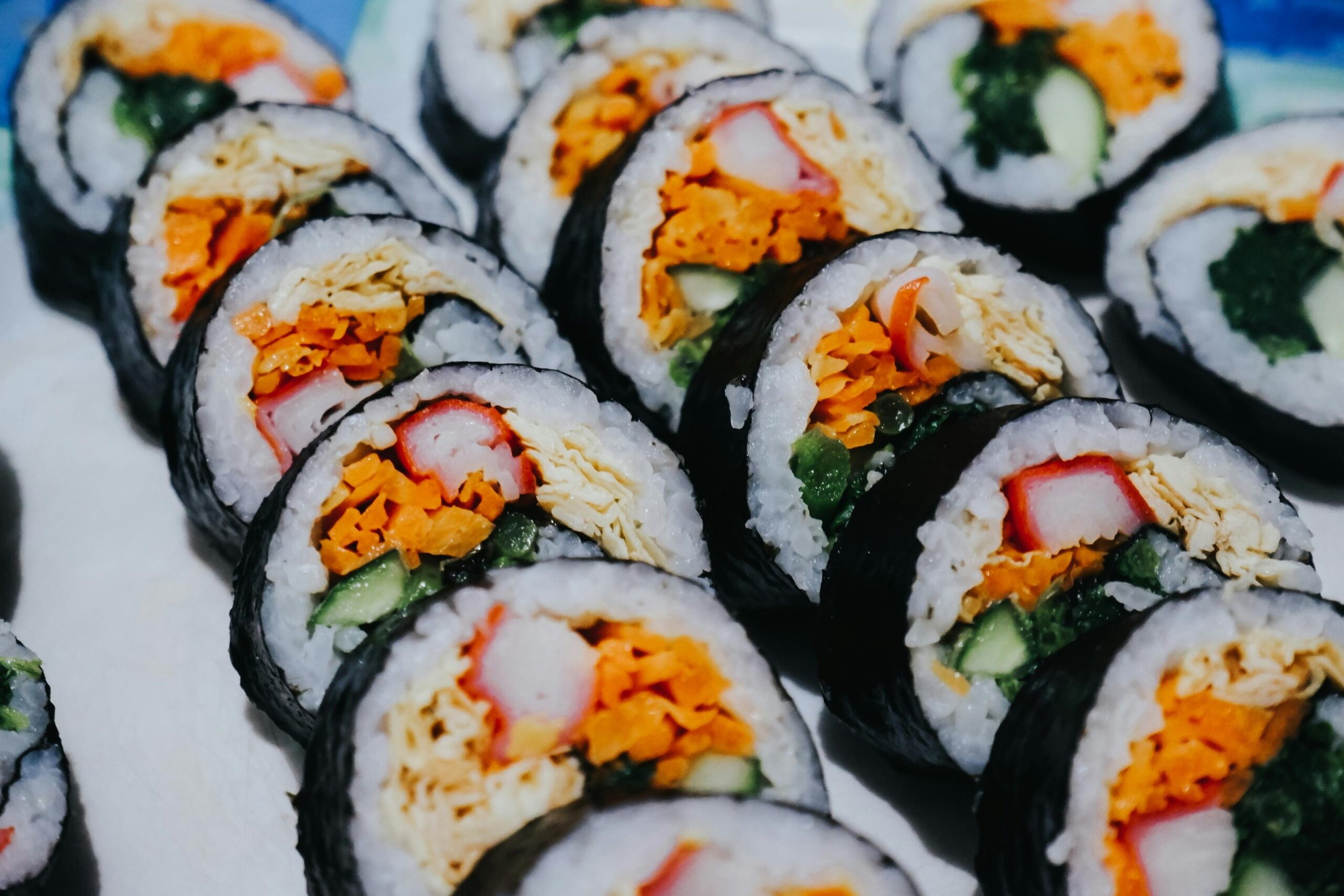 This close-up view presents diagonal rows of sliced sushi rolls, featuring a core of colorful ingredients tightly wrapped in white rice and dried seaweed. The fillings include vibrant strips of crab meat, orange carrots, yellow egg, and green vegetables, creating a visually appetizing and textured pattern.