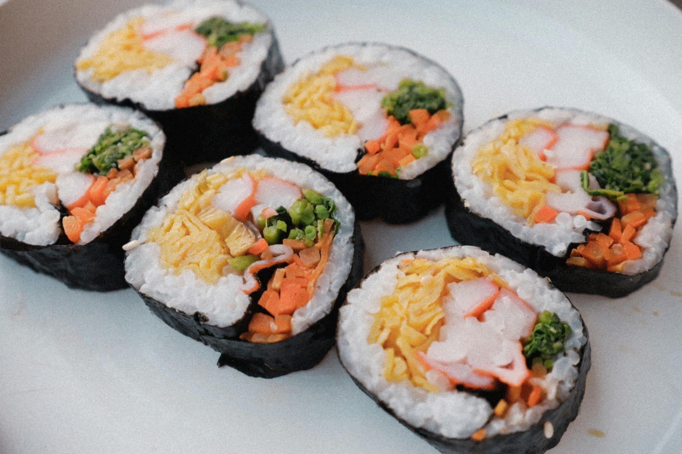 This close-up shot features a small group of sliced sushi rolls arranged on a white plate, highlighting their vibrant fillings. Wrapped in black seaweed and white rice, the rolls contain distinct sections of yellow egg, orange carrots, crab stick, and green vegetables.