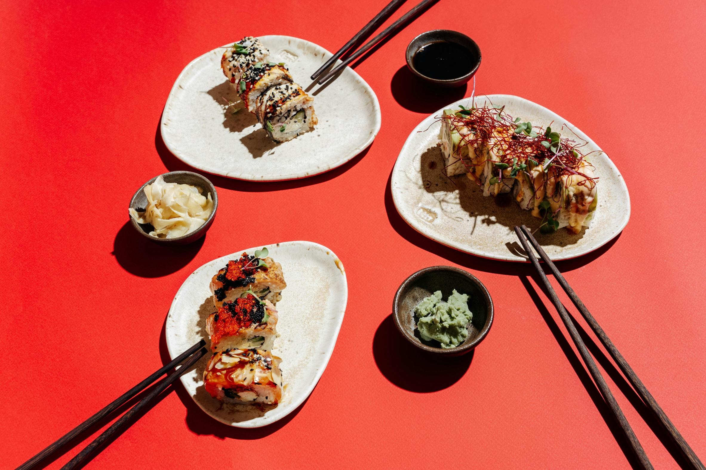 This overhead flat-lay composition presents three rustic plates of diverse sushi rolls, garnished with toppings like sesame seeds, roe, and chili threads, set against a bold red background. Dark wooden chopsticks and small bowls filled with soy sauce, wasabi, and pickled ginger surround the main dishes.