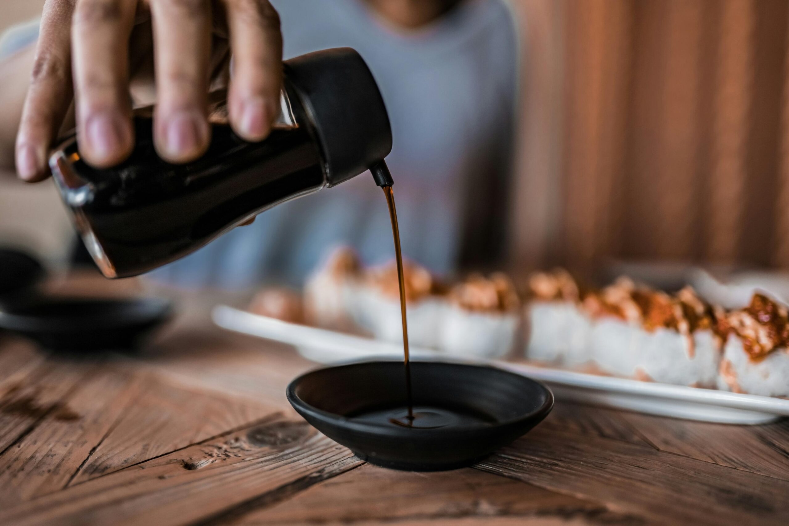 A hand pours dark soy sauce from a glass dispenser into a small black dipping bowl. In the blurred background, a plate of sushi rolls rests on a rustic wooden table, ready to be eaten.