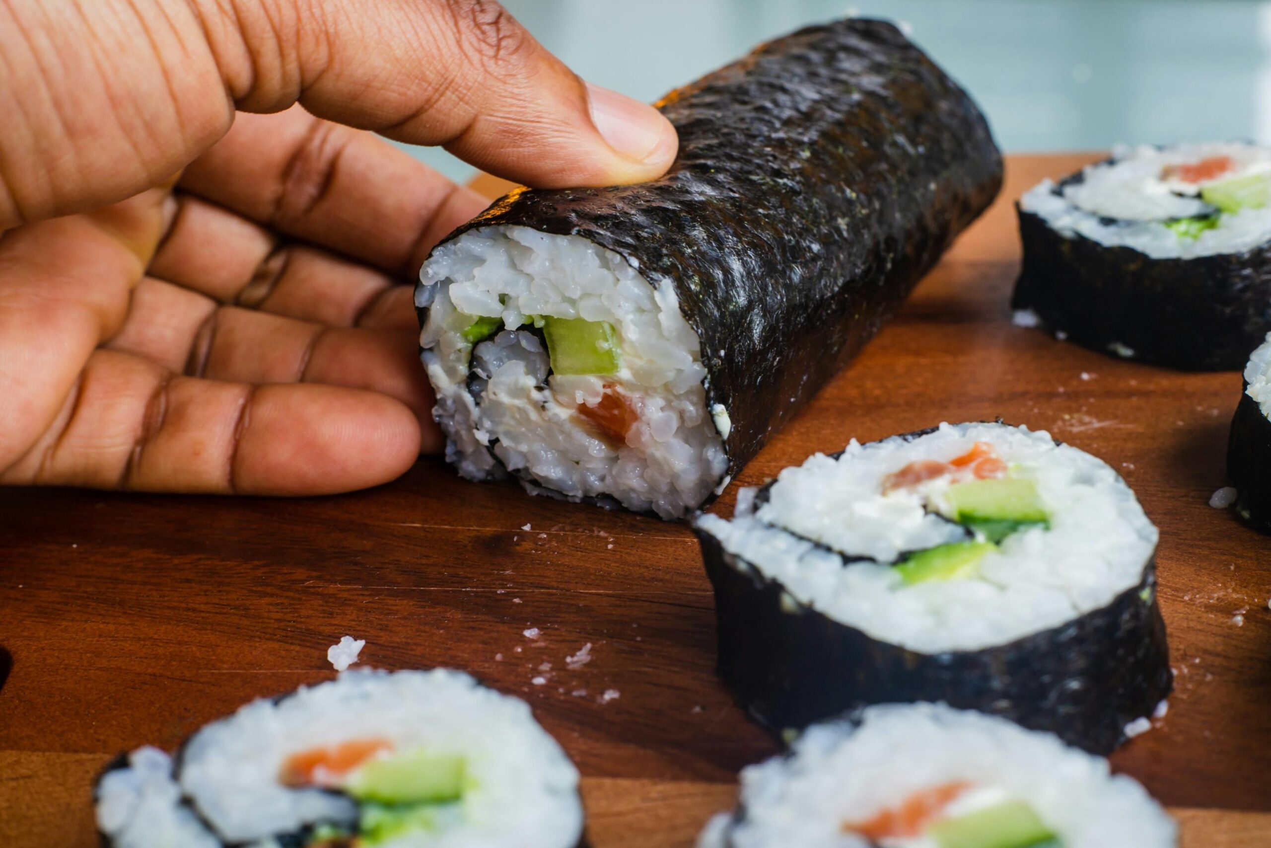 A close-up captures a hand holding a thick, unsliced portion of a nori-wrapped sushi roll on a wooden surface. Surrounding the roll are several cut segments displaying a filling of white rice, salmon, cream cheese, and cucumber.