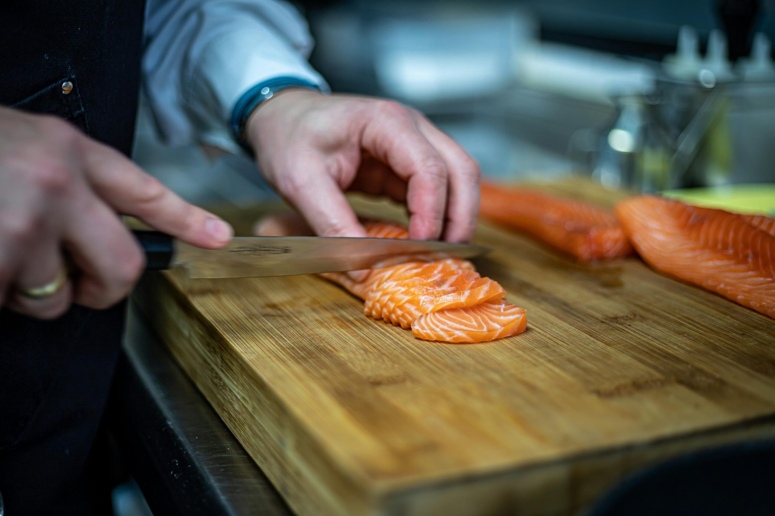 A close-up shot captures the hands of a chef skillfully slicing fresh, raw salmon on a sturdy wooden cutting board. With a large fillet waiting in the background, the scene highlights the precise preparation involved in making sushi or sashimi.
