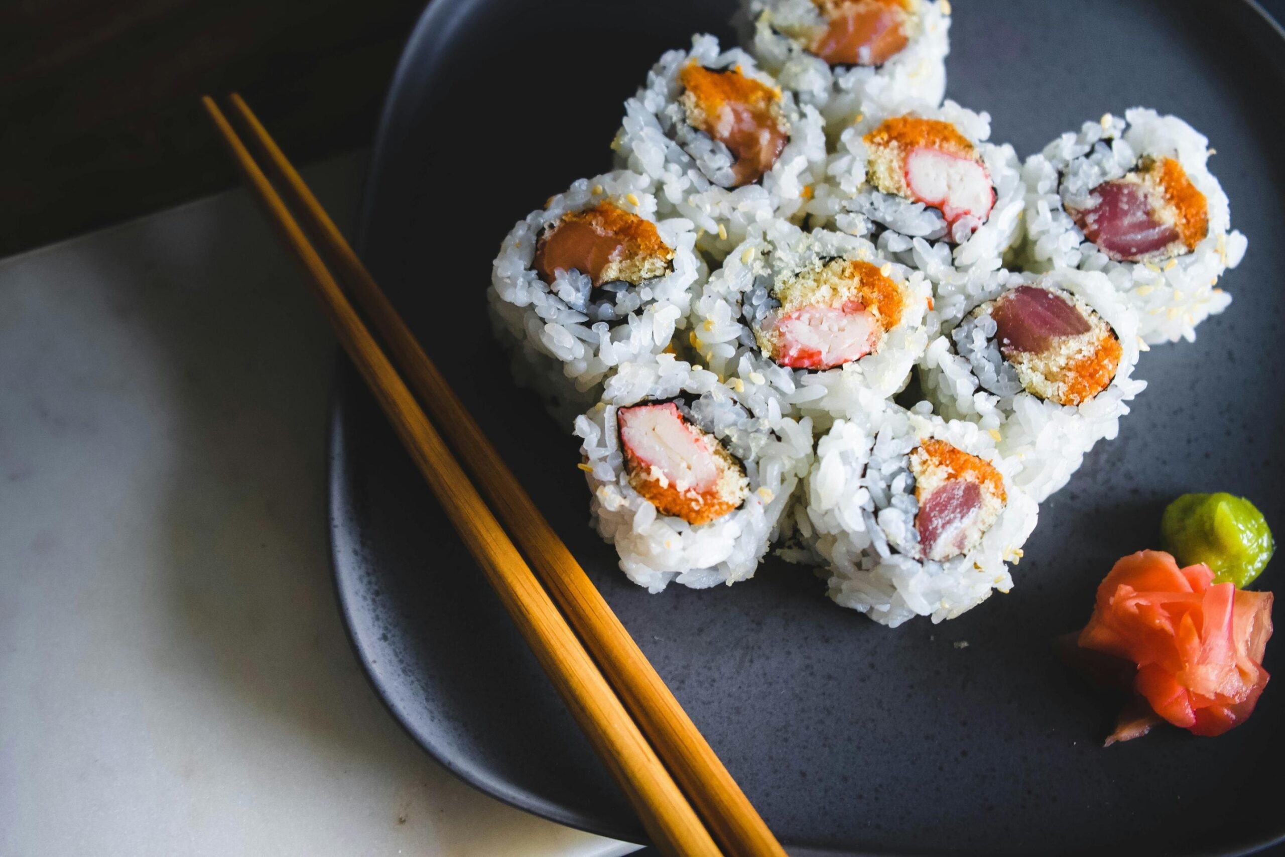 This overhead shot features a cluster of inside-out sushi rolls filled with crab and tuna, sprinkled with sesame seeds and arranged on a dark grey plate. To complete the serving, a pair of wooden chopsticks rests on the left, accompanied by a small dollop of green wasabi and pink pickled ginger on the right.