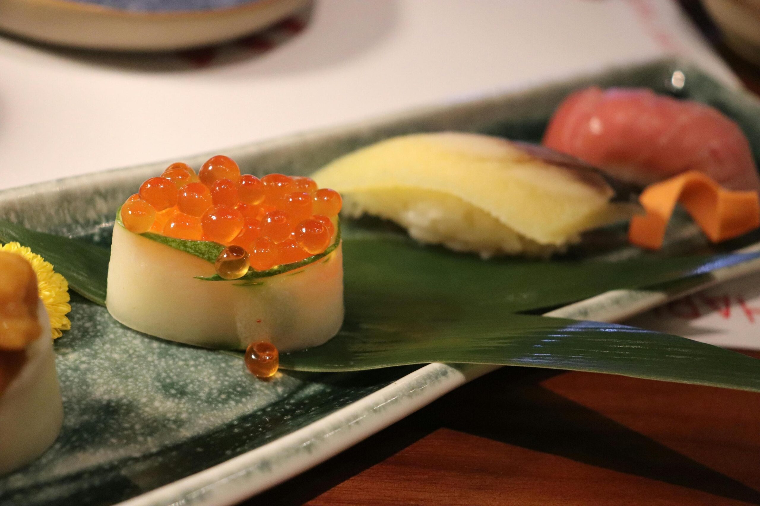 This close-up captures a piece of sushi topped with glistening orange roe and wrapped in a slice of cucumber, sitting on a glazed rectangular plate. In the background, other nigiri pieces rest on a bamboo leaf garnish, completing the elegant Japanese food presentation.