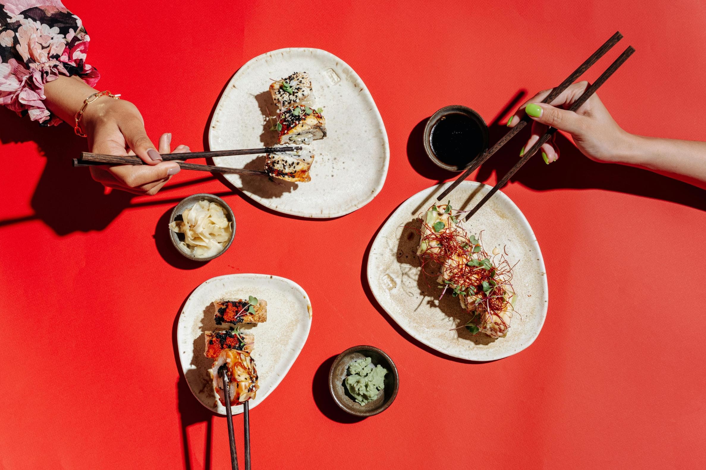 This overhead composition shows two people sharing a meal, using chopsticks to pick from an assortment of sushi rolls garnished with toppings like chili threads and sesame seeds. The scene features a bold red background where direct, hard lighting creates sharp shadows around the rustic plates and condiment bowls containing soy sauce, wasabi, and ginger.