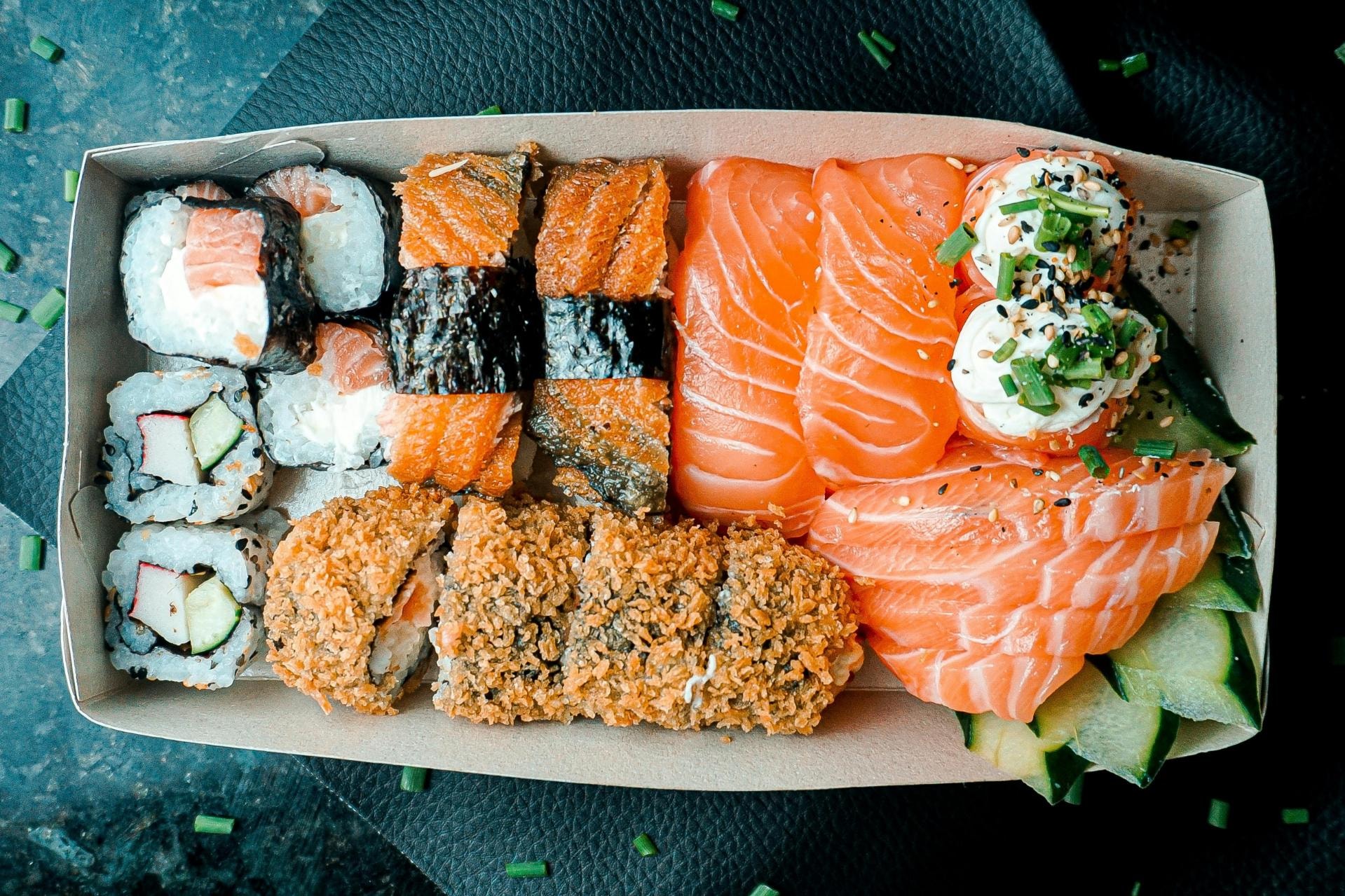 This overhead shot captures a cardboard takeout box packed with a delicious assortment of sushi, including fresh salmon nigiri, sashimi, maki, and crispy fried rolls. The colorful arrangement features varied textures and toppings, such as cream cheese and sesame seeds, set against a dark background sprinkled with chopped green onions.
