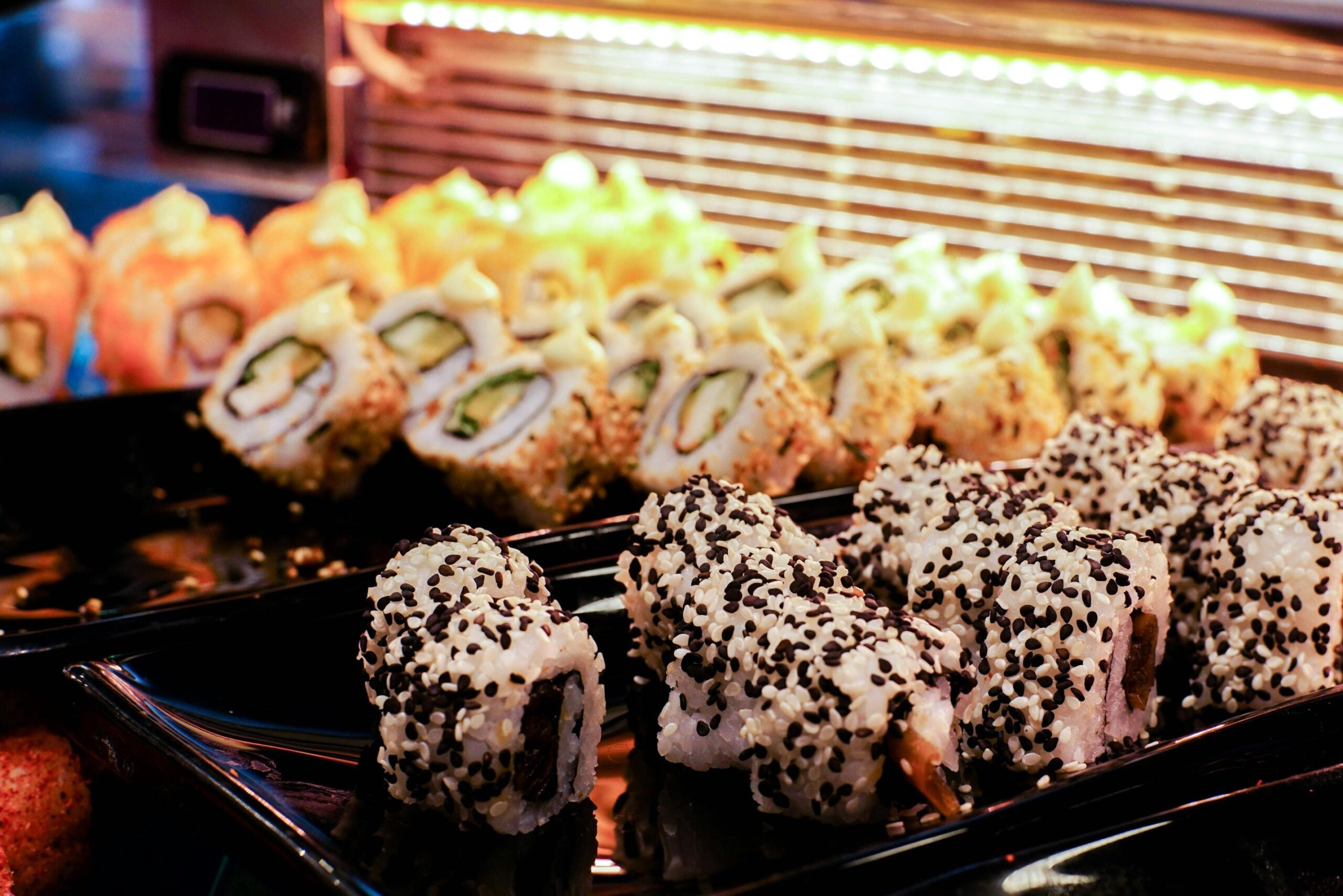 This image captures a vibrant display of various sushi rolls arranged on black trays within a refrigerated glass case. The assortment features rolls coated in black and white sesame seeds in the foreground, with others topped with creamy sauces and colorful fillings receding into the warm, glowing background.