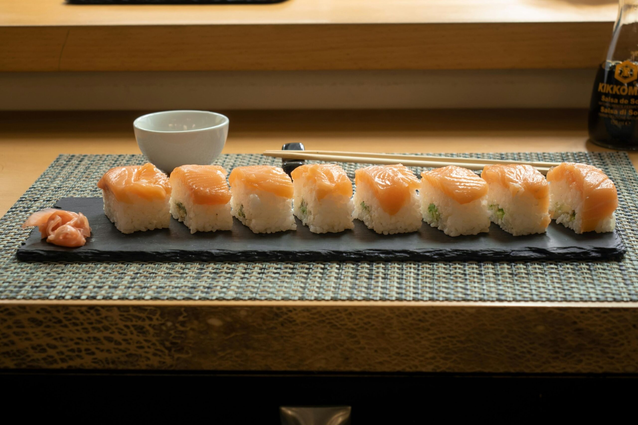 This image features a neat row of eight salmon sushi pieces served on a dark slate platter alongside a small portion of pickled ginger. The set is neatly arranged on a textured placemat with chopsticks, a small white bowl, and a bottle of soy sauce in the background.
