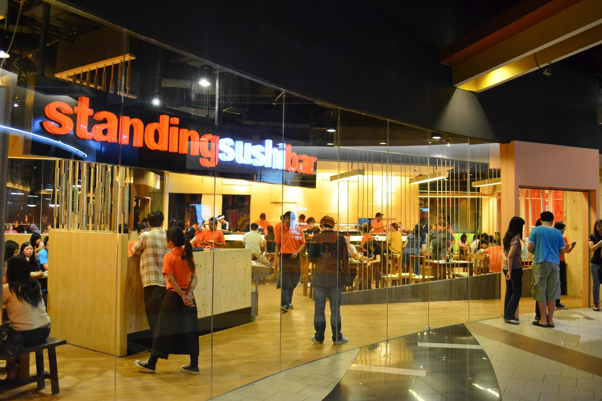 The Standing Sushi Bar features a large glass storefront with glowing red and white signage overlooking a busy interior where staff in orange shirts serve customers. Inside, the warm-toned space is filled with people dining at wooden tables and counters beneath modern light fixtures.