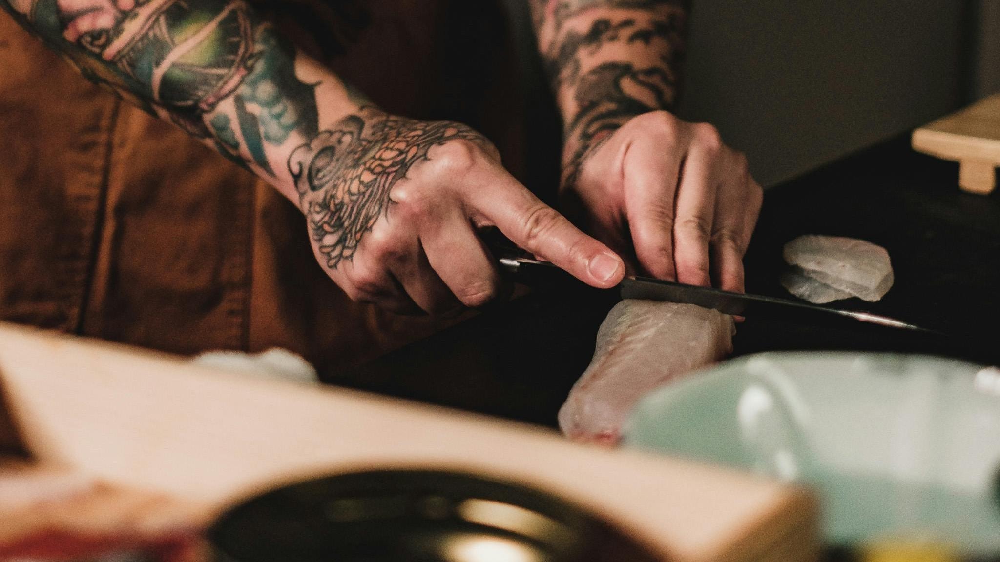 A chef with tattooed arms carefully slices a piece of raw white fish on a dark surface using a sharp thin knife.
