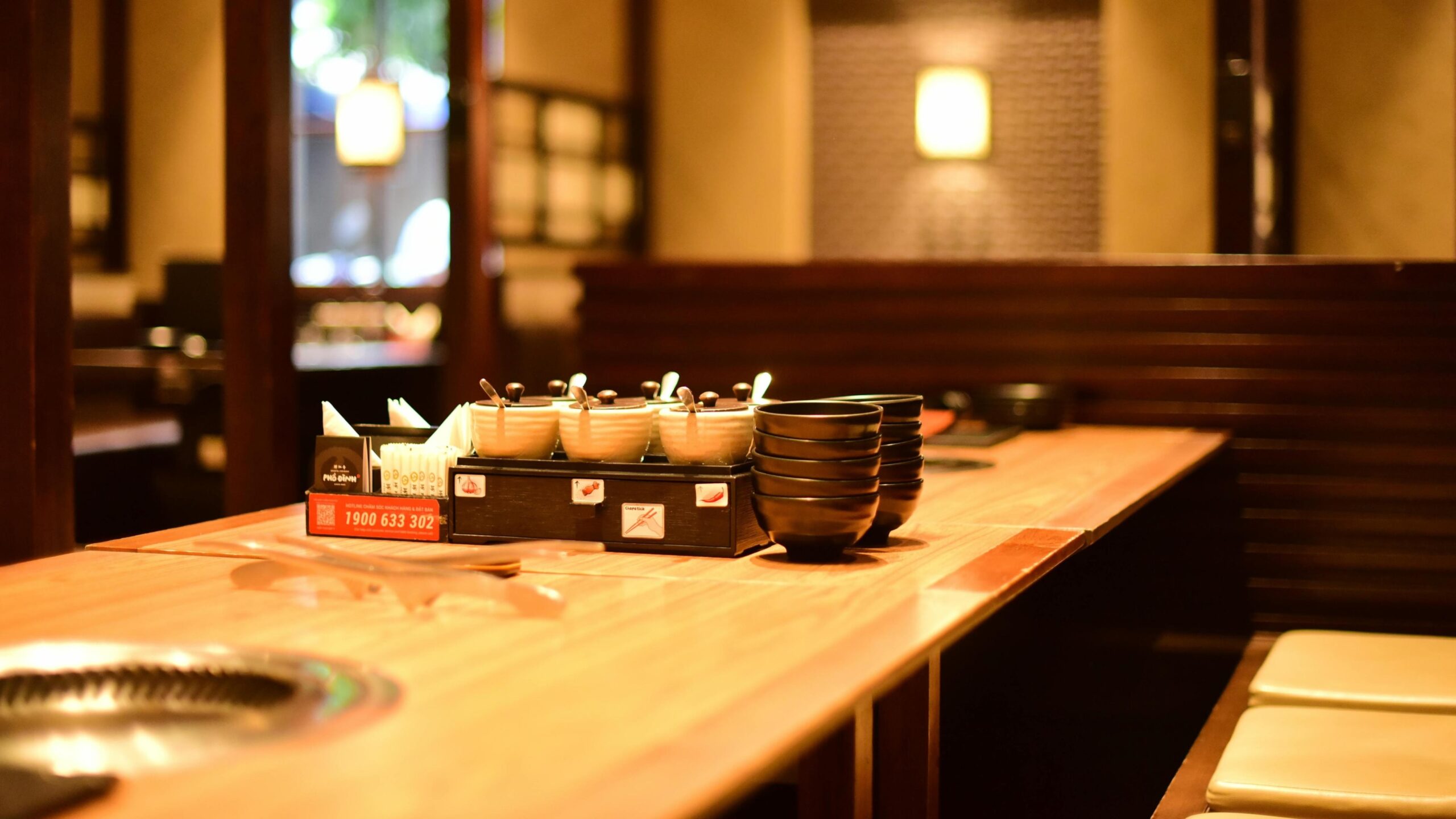 In a warm, wood-toned restaurant, a dining table is neatly organized with stacks of black bowls, small condiment jars, and a recessed grill for tabletop cooking.