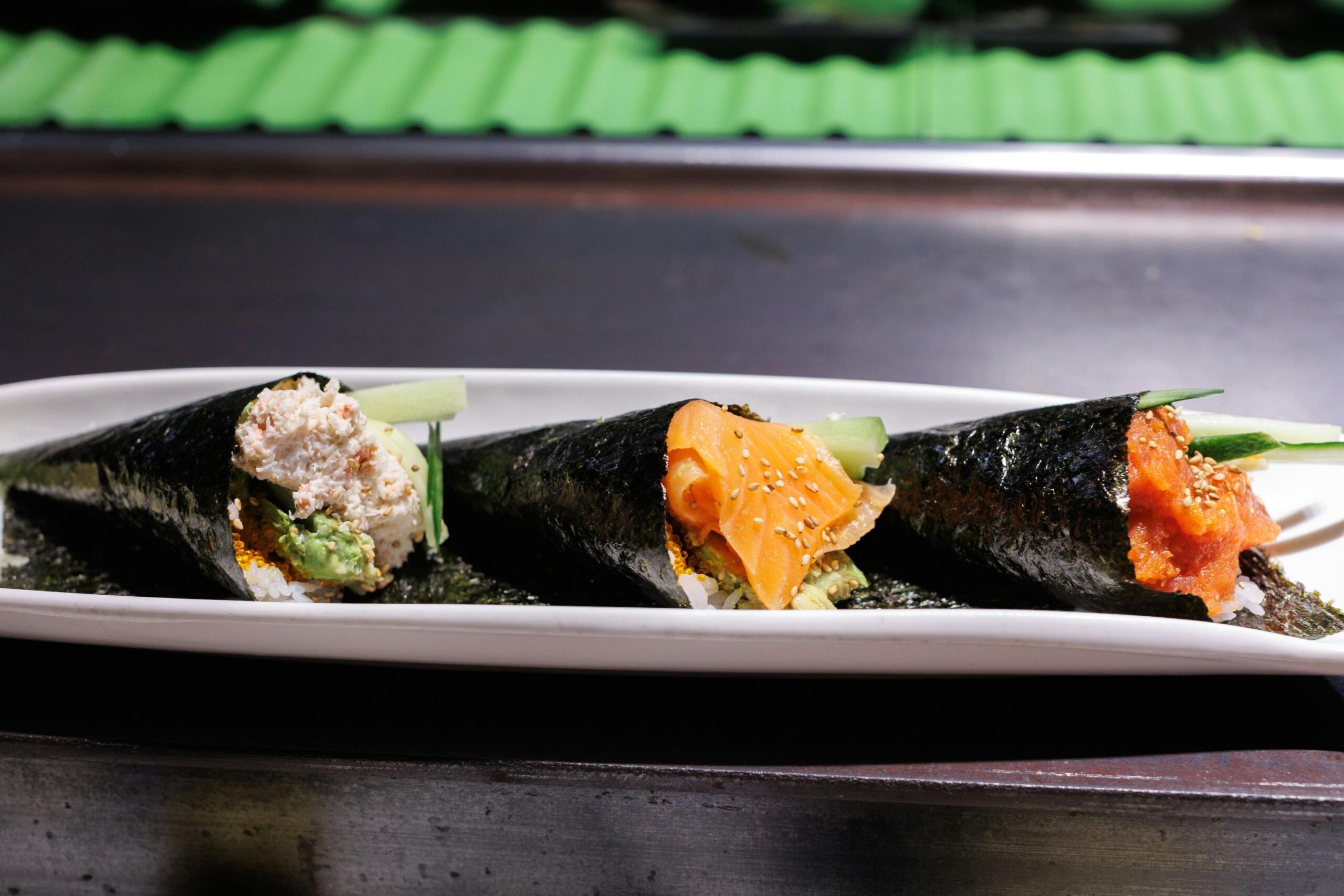  Three hand-rolled temaki sushi cones filled with various ingredients like fish, cucumber, and avocado are arranged on a long white platter. The rolls are displayed in a commercial setting, resting on a dark surface with a textured green background visible behind them.