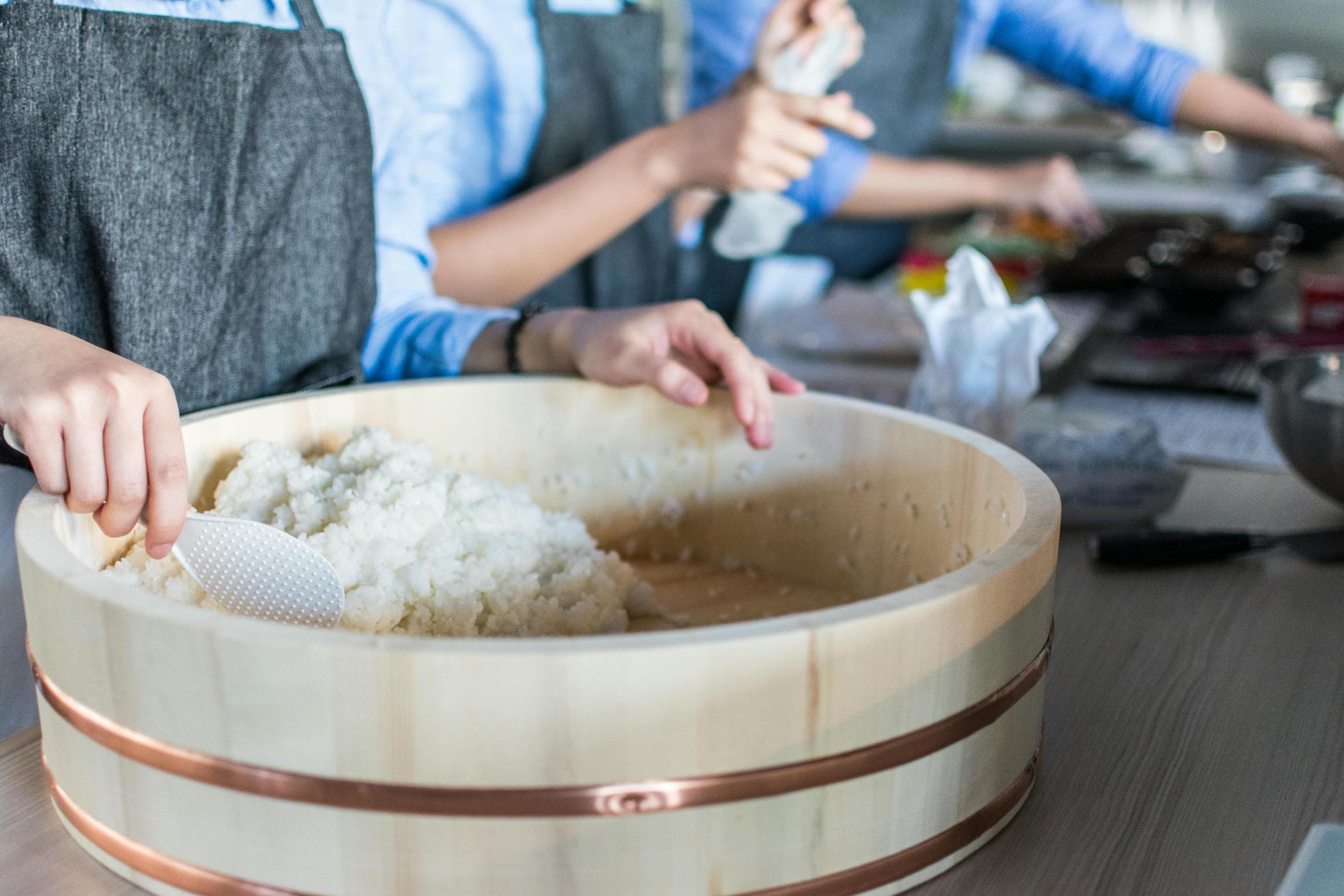 A person wearing a dark apron uses a white paddle to mix cooked rice inside a large wooden tub. In the background, other individuals are also engaged in food preparation tasks along a counter.