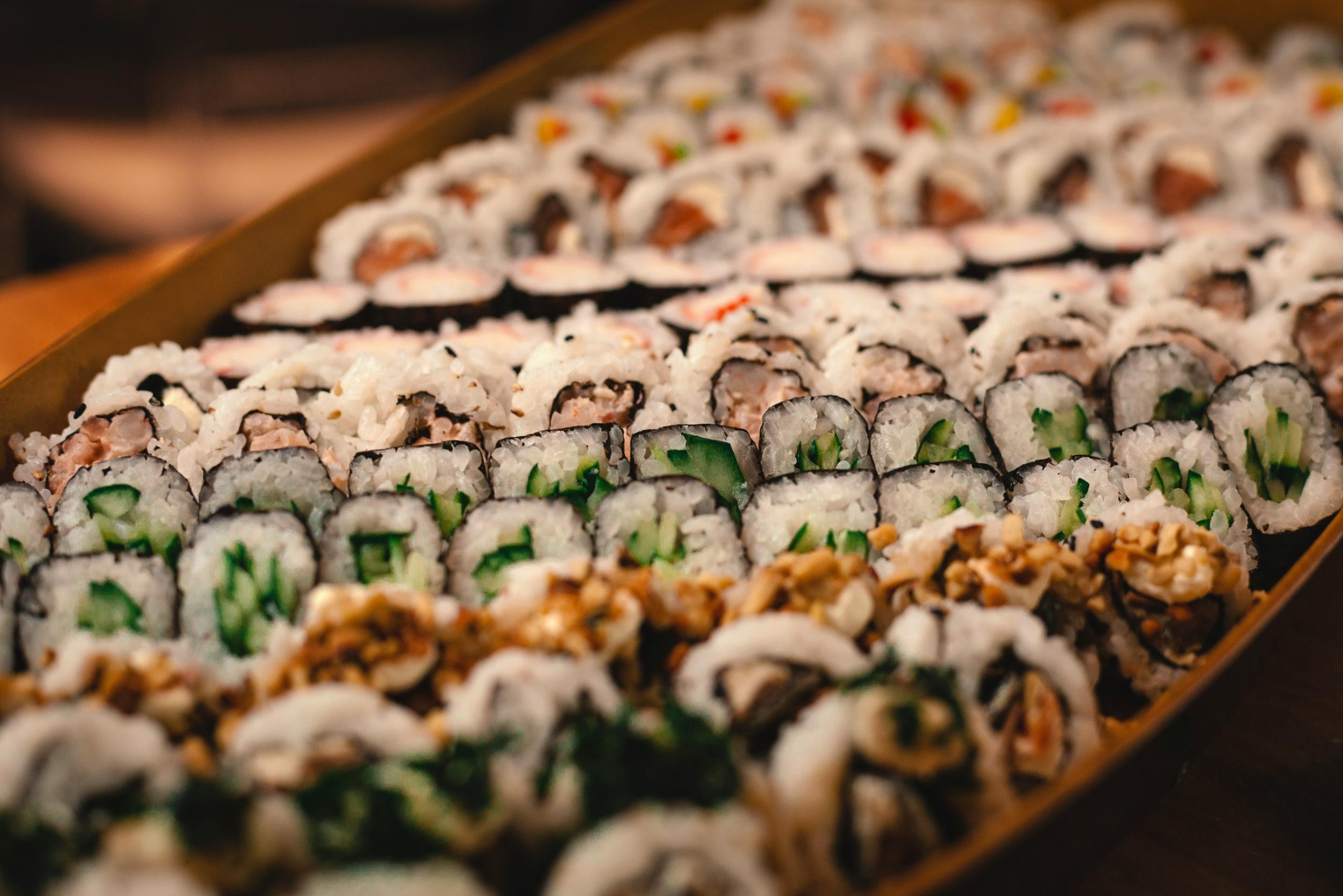  A large, rectangular wooden tray is densely packed with rows of diverse sushi rolls, featuring various fillings like cucumber, salmon, and tempura. The image uses a shallow depth of field to focus on the textures of the middle rows while the front and back of the assortment softly blur.