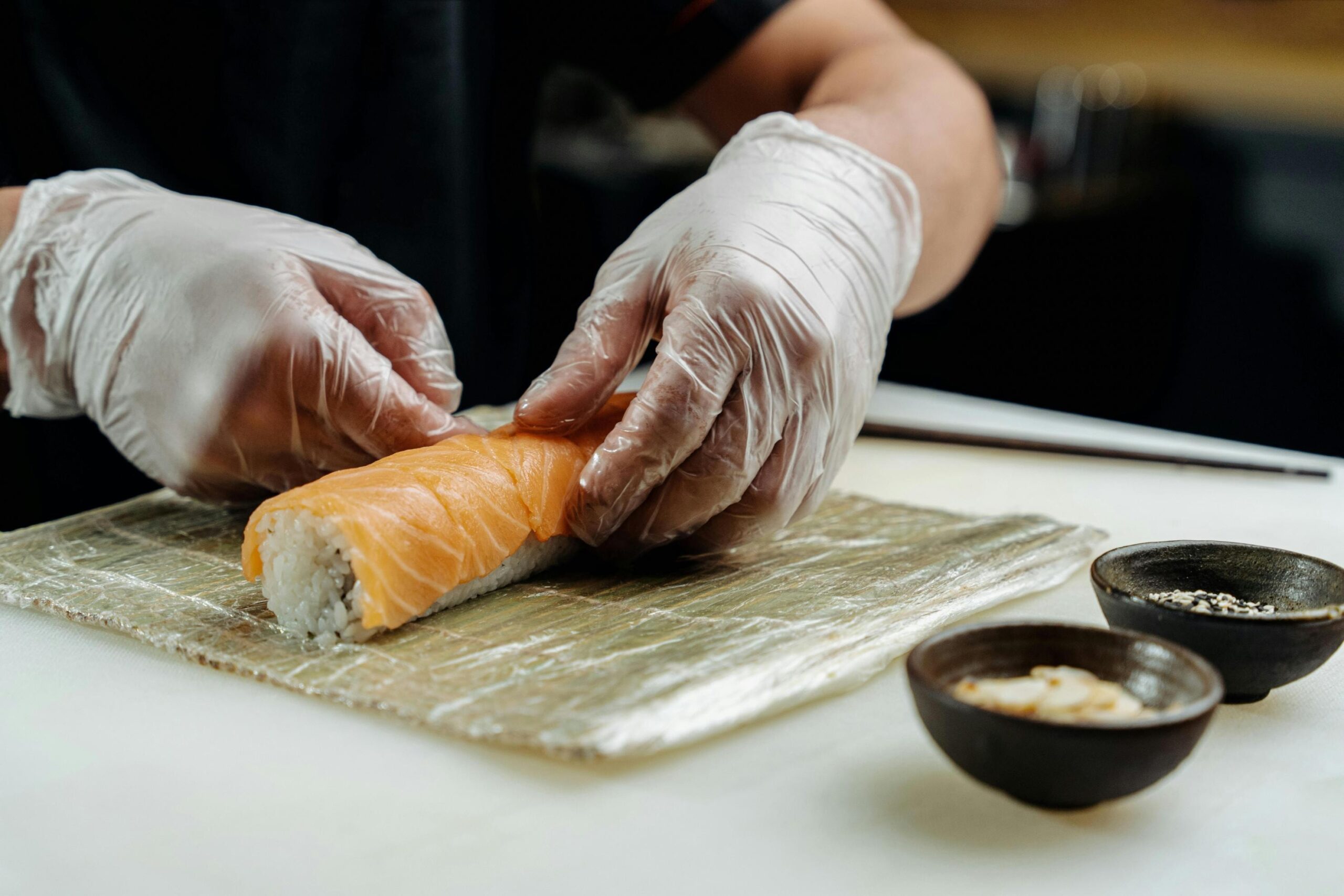 A chef wearing clear gloves carefully shapes a sushi roll topped with fresh salmon on a bamboo mat. Two small bowls containing garnishes sit nearby on the clean workspace.