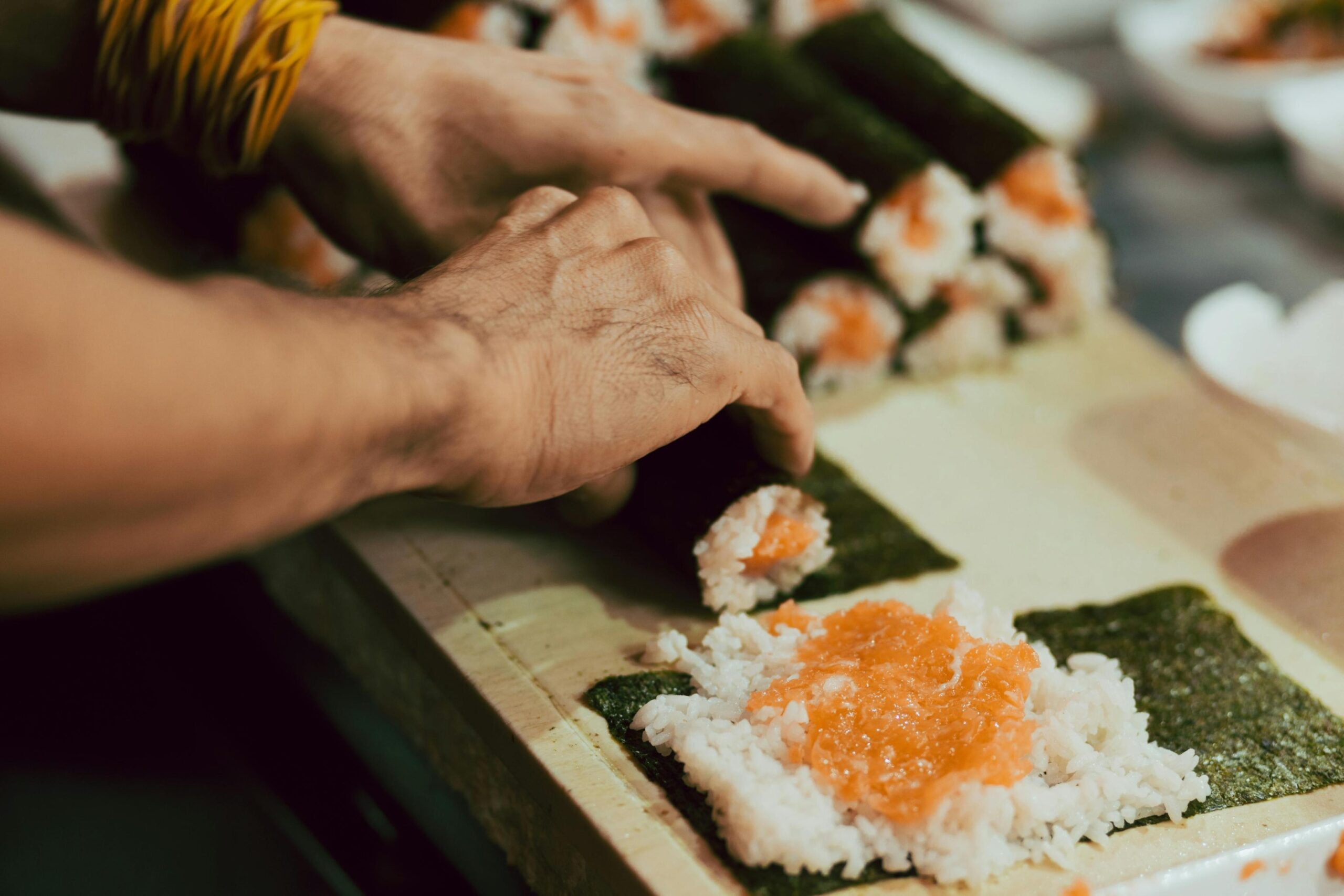 Hands expertly roll sushi using sheets of nori topped with white rice and orange fish. A finished stack of these hand-rolled sushi cones sits in the background on the prep surface.