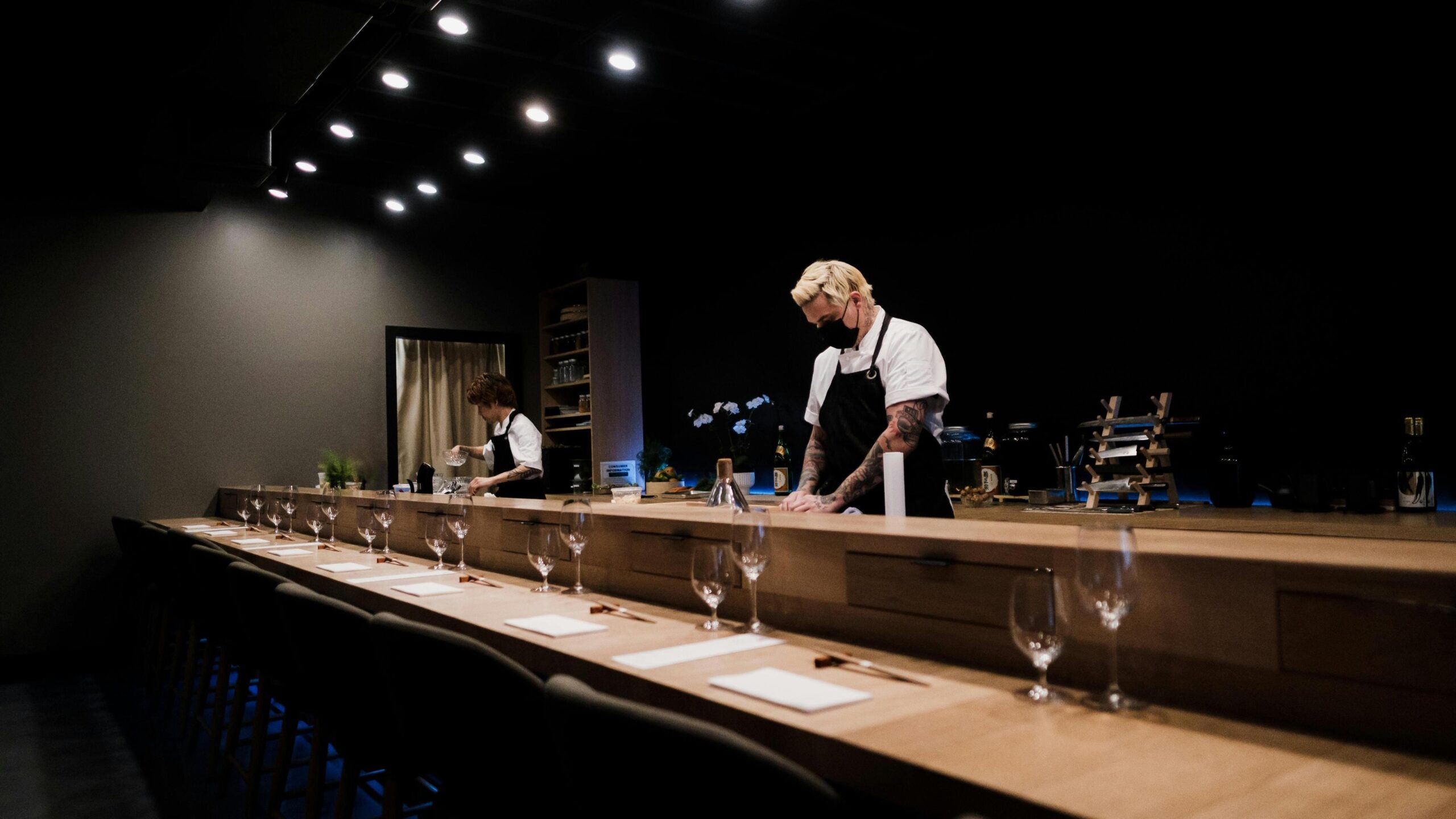 In a sleek, dimly lit modern restaurant, two chefs prepare food behind a long wooden counter meticulously set with glassware and napkins for an upscale dining experience.