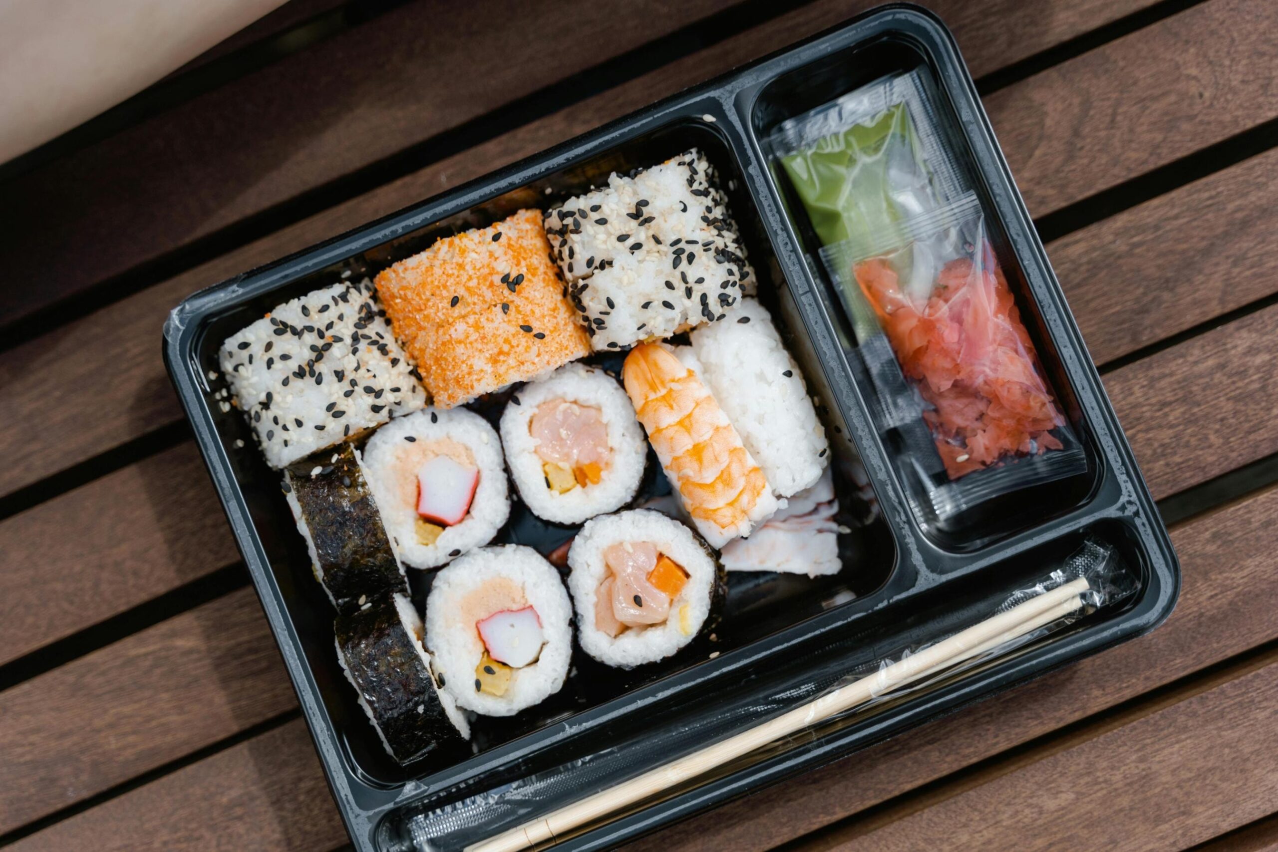 An overhead view shows a black bento box filled with an assortment of sushi rolls and nigiri resting on a wooden slatted surface. The container includes separate compartments for chopsticks and small packets of ginger and wasabi.